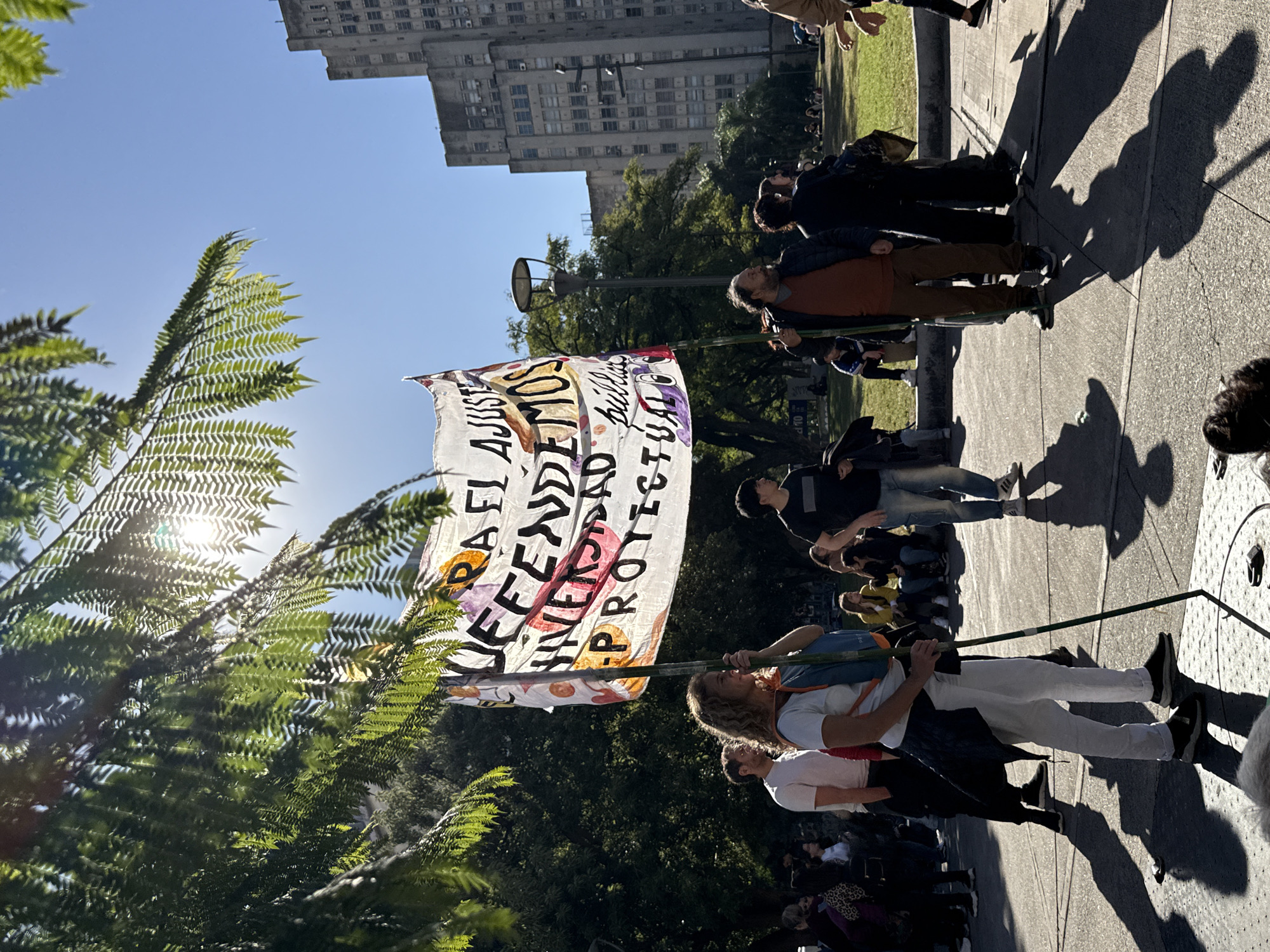 A daytime shot captures a protest in what is likely Buenos Aires, Argentina. People are assembled on a paved area lined by green spaces and trees, with a tall building in the background. The focus is on a white banner with the words "DEFENDA EL AJUSTE", "LIBERTAD", and "PROTECT" written on it in Spanish and English. Two individuals hold the banner aloft. Various individuals are visible in the crowd, with shadows cast on the ground by the sun. A palm tree is visible to the left of the banner.