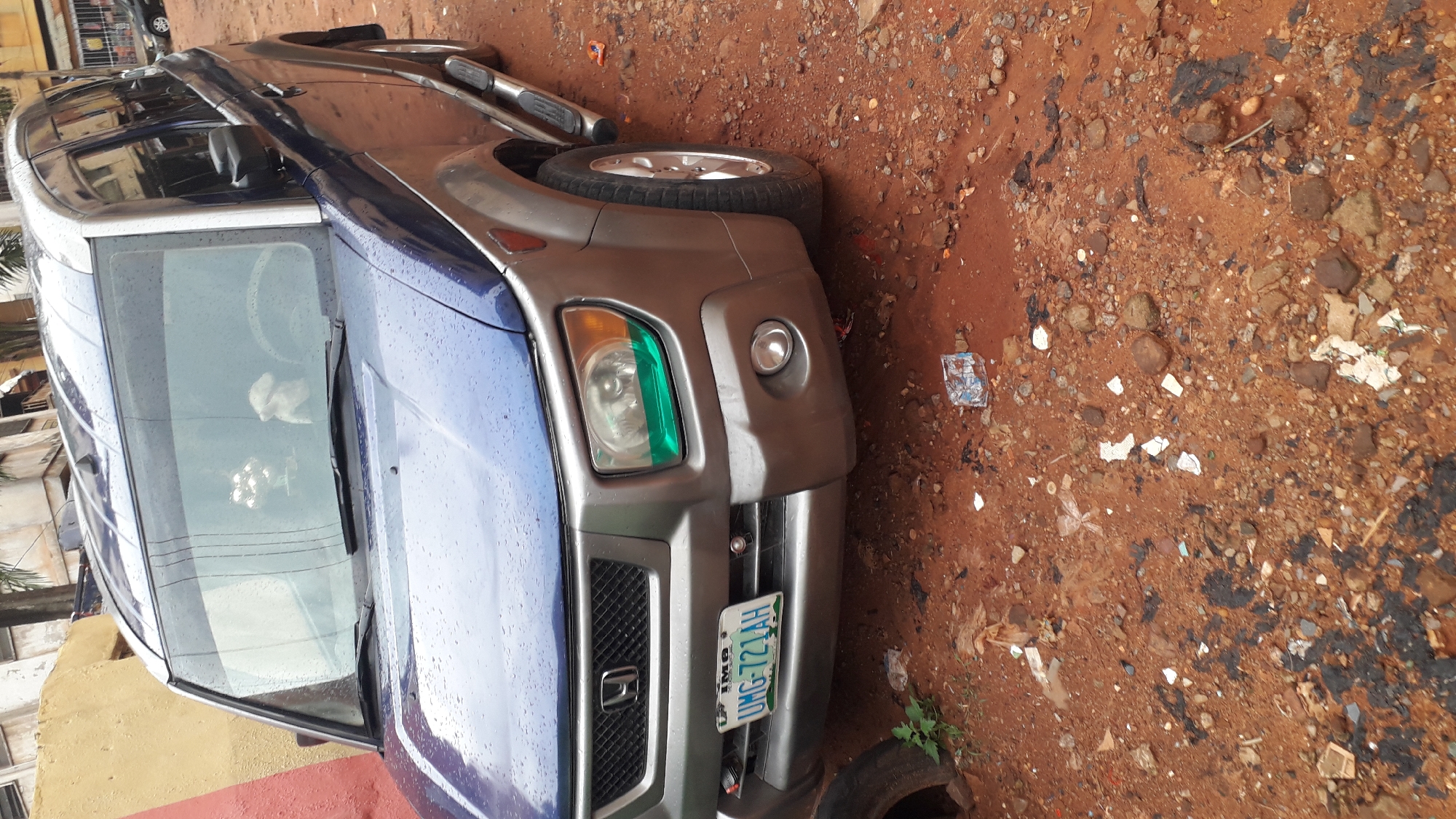 A blue and gray Honda Element is parked on a dirt road in Onitsha, Nigeria. The vehicle has a Nigerian license plate that reads UMG-721AH. The area surrounding the car is littered with trash and debris.