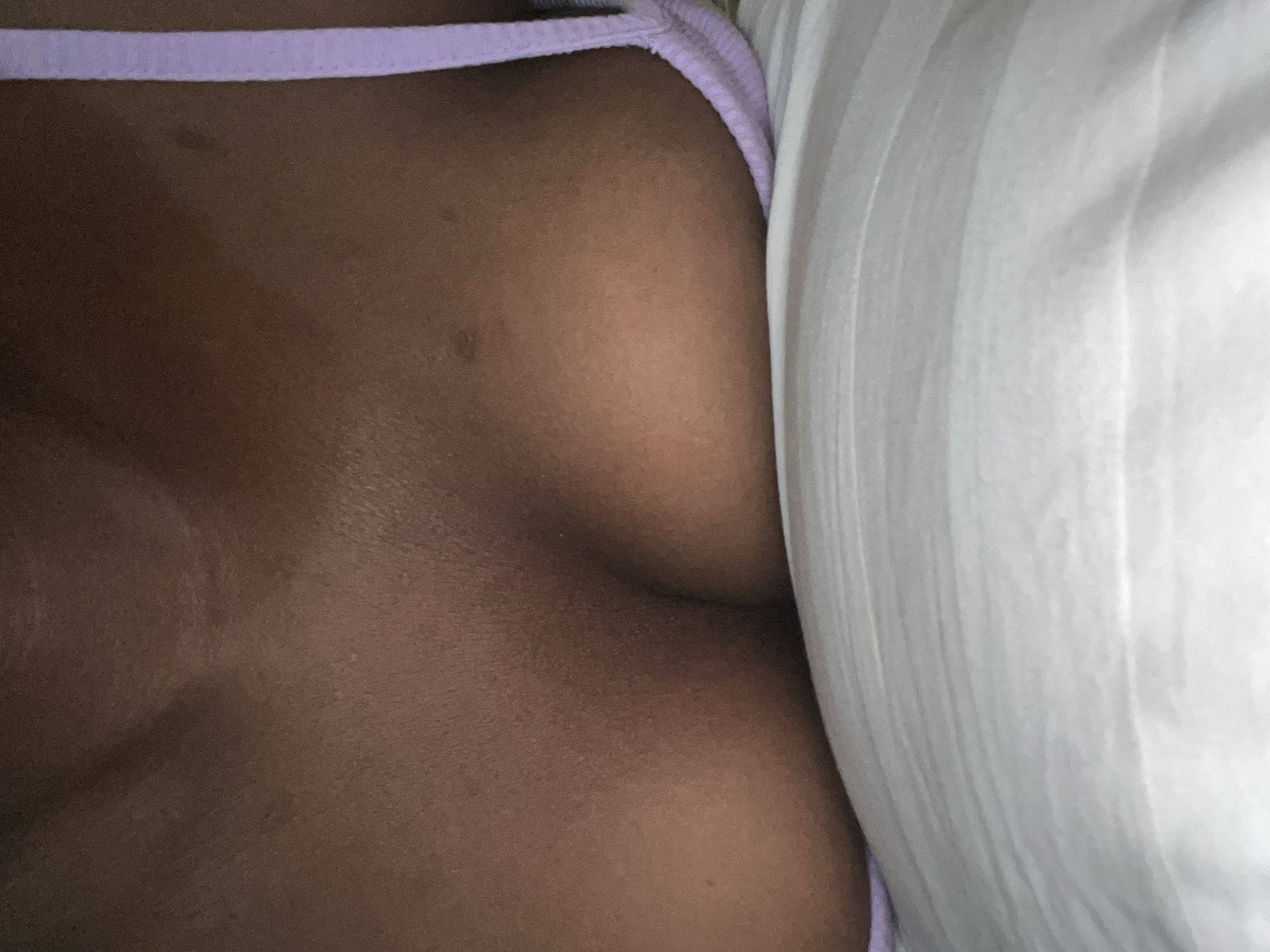 The image is a close-up, chest-level shot of a person lying down on a white pillow. The person is wearing a light purple bra strap visible on the right side of the frame. The skin tone is dark. The lighting appears to be natural, coming from the top. The background is out of focus, showing a soft texture of what seems to be a bedsheet or pillow. No specific details about the location in Howayi, Nigeria can be deduced from the image.