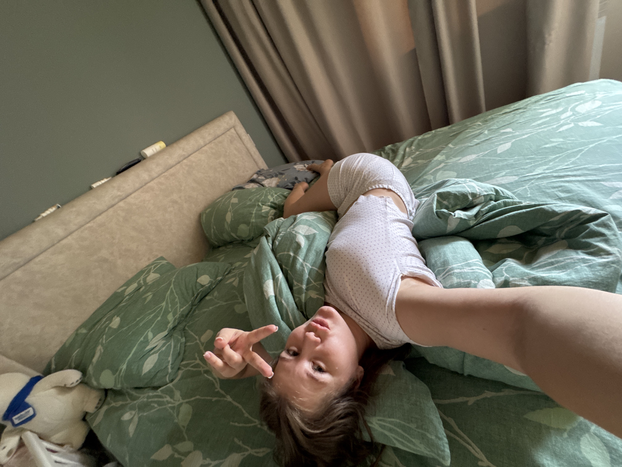A young woman is lying upside down on a bed with green leaf-patterned bedding. She is wearing a white polka-dot top and shorts and is making a peace sign with her left hand. In the background, there is a beige headboard and curtains. A stuffed animal is located on the lower left corner. The image was taken in Ферма, Russia.