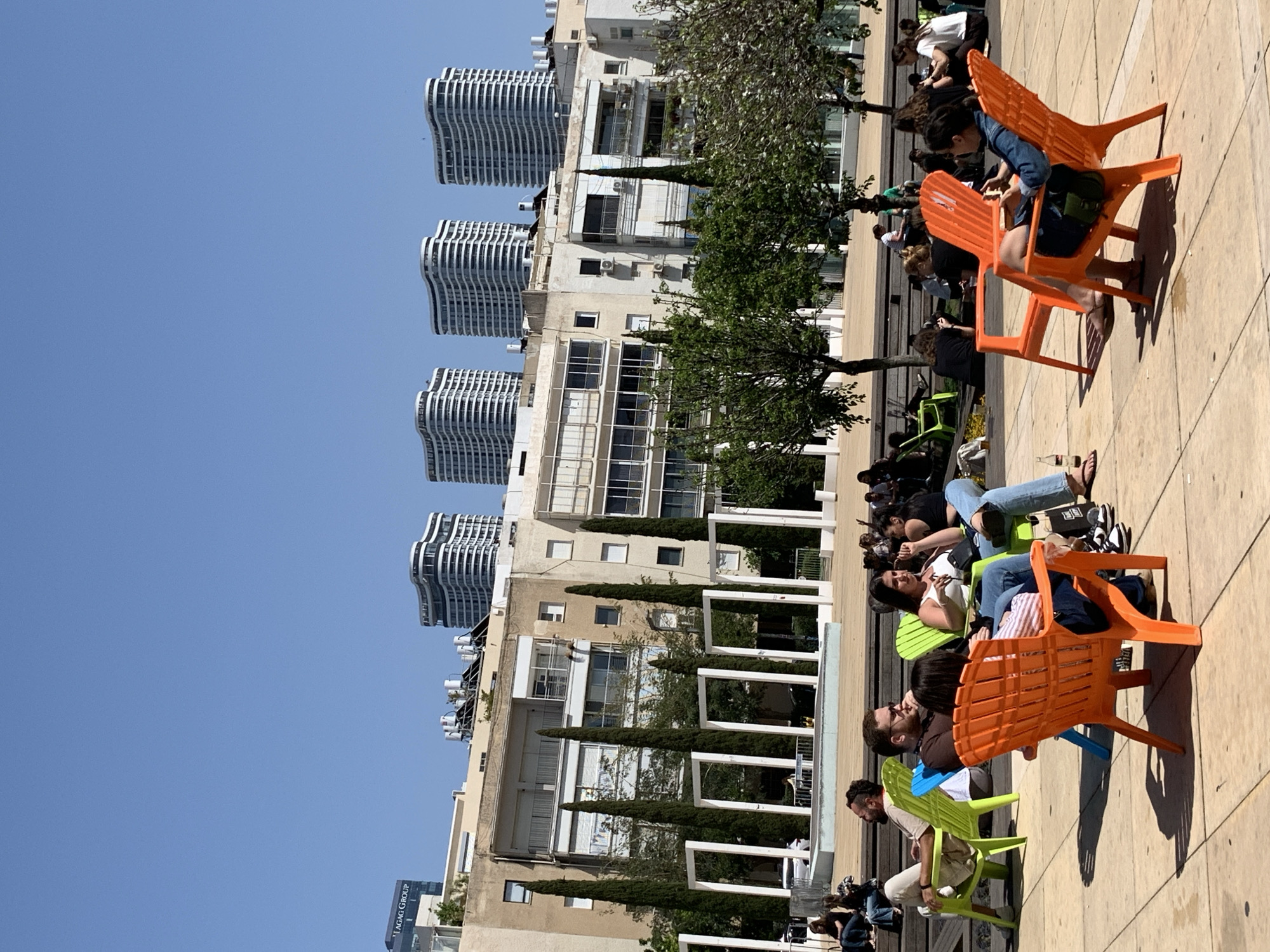 The image shows a sunny scene in Tel-Aviv, Israel, with modern architecture and a group of people relaxing outdoors. In the background, distinctive wavy, white high-rise buildings are visible against a clear blue sky, a characteristic of Tel-Aviv's urban landscape. Closer to the foreground are older, lower-rise buildings with balconies, trees, and decorative white structures. The primary focus is on a group of people lounging in bright orange and green plastic chairs on a paved surface, likely a public square or outdoor seating area. The people are dressed casually and appear to be conversing or relaxing. Some are facing forward, while others are turned to the side. Some shadows are visible on the ground, which indicates that it is daytime and the sun is shining. The overall impression is one of urban leisure and relaxation within the modern cityscape of Tel-Aviv.