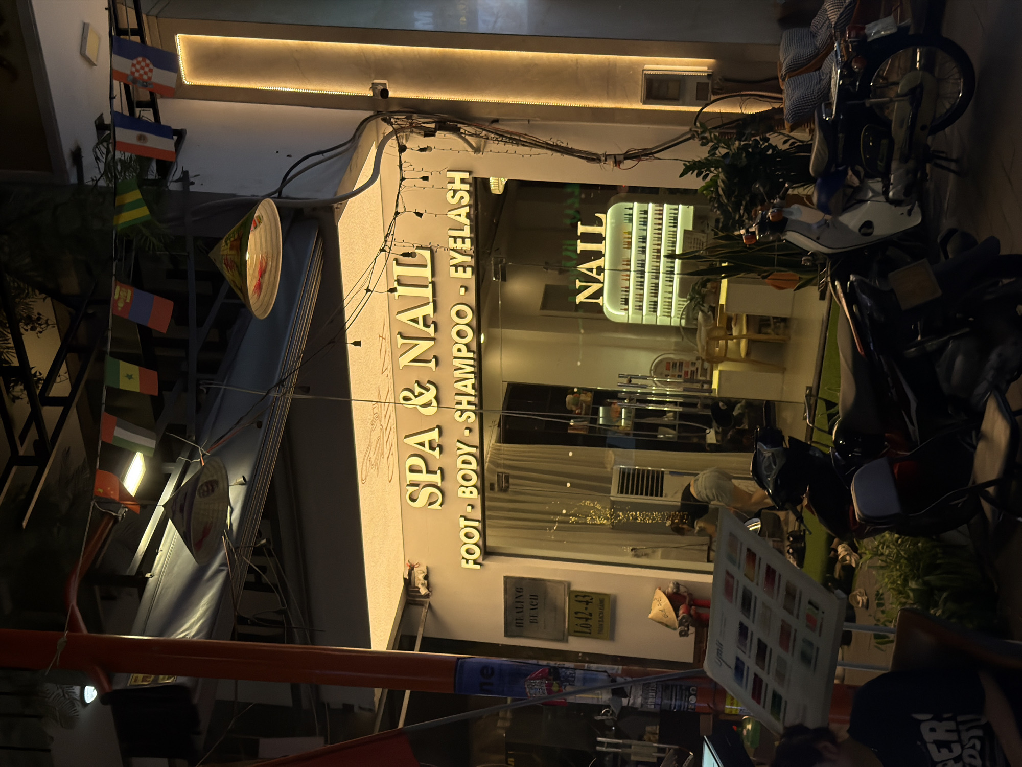The image depicts a nighttime street scene in Da Nang, Vietnam, featuring a brightly lit spa and nail salon. The salon's facade is illuminated by warm, yellow lighting, with prominent signage that reads "SPA & NAIL" in large, gold-colored lettering, followed by smaller text listing services: "FOOT - BODY - SHAMPOO - EYELASH". Below this, another illuminated sign reads "NAIL" in a modern font, above a display case filled with rows of nail polish bottles.
Several motorcycles are parked in front of the establishment, indicating a common mode of transport in the area. To the left of the salon entrance, a menu board displays various services with colored swatches, likely for nail polish or treatments. A person is visible inside the salon, appearing to be interacting with the display or a customer. Outside, a woman in casual attire is seen in the background near the motorcycles.
The environment suggests a commercial street with a slightly busy atmosphere. Decorative flags from various countries are strung across the upper part of the scene, and a traditional conical hat is placed as an ornament near a red post. The overall impression is that of a well-maintained business catering to beauty and relaxation services, typical of an urban setting in Vietnam.
