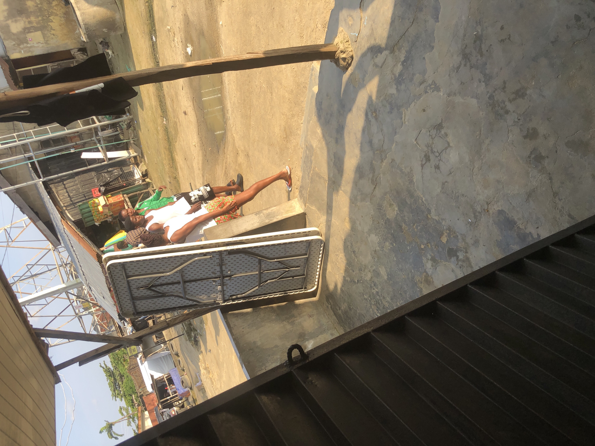 In the city of Ado, Nigeria, a scene unfolds outdoors under a bright sky, characterized by a dusty, unpaved ground scattered with puddles. In the foreground, a set of dark, slatted stairs descends from the left. To the right of the stairs, a folded white plastic table stands upright against a low concrete wall. Beyond the wall, the ground is a mix of sand and dry earth.
Three individuals are present. The first, a woman, is leaning against the concrete wall, her body angled away from the viewer. She is wearing a white top and colorful patterned shorts. A second individual, a man, stands to her left, wearing a white tank top and black and white patterned shorts. A third man stands further left, facing the viewer, dressed in a green jacket and dark shorts. Behind them, a makeshift structure with a corrugated metal roof is visible, along with various objects and debris, including crates and what appear to be tires stacked. Further in the background, a tall metal structure with scaffolding is visible. Chickens peck at the ground in the middle ground. The overall impression is one of an outdoor market or residential area in a developing urban environment.