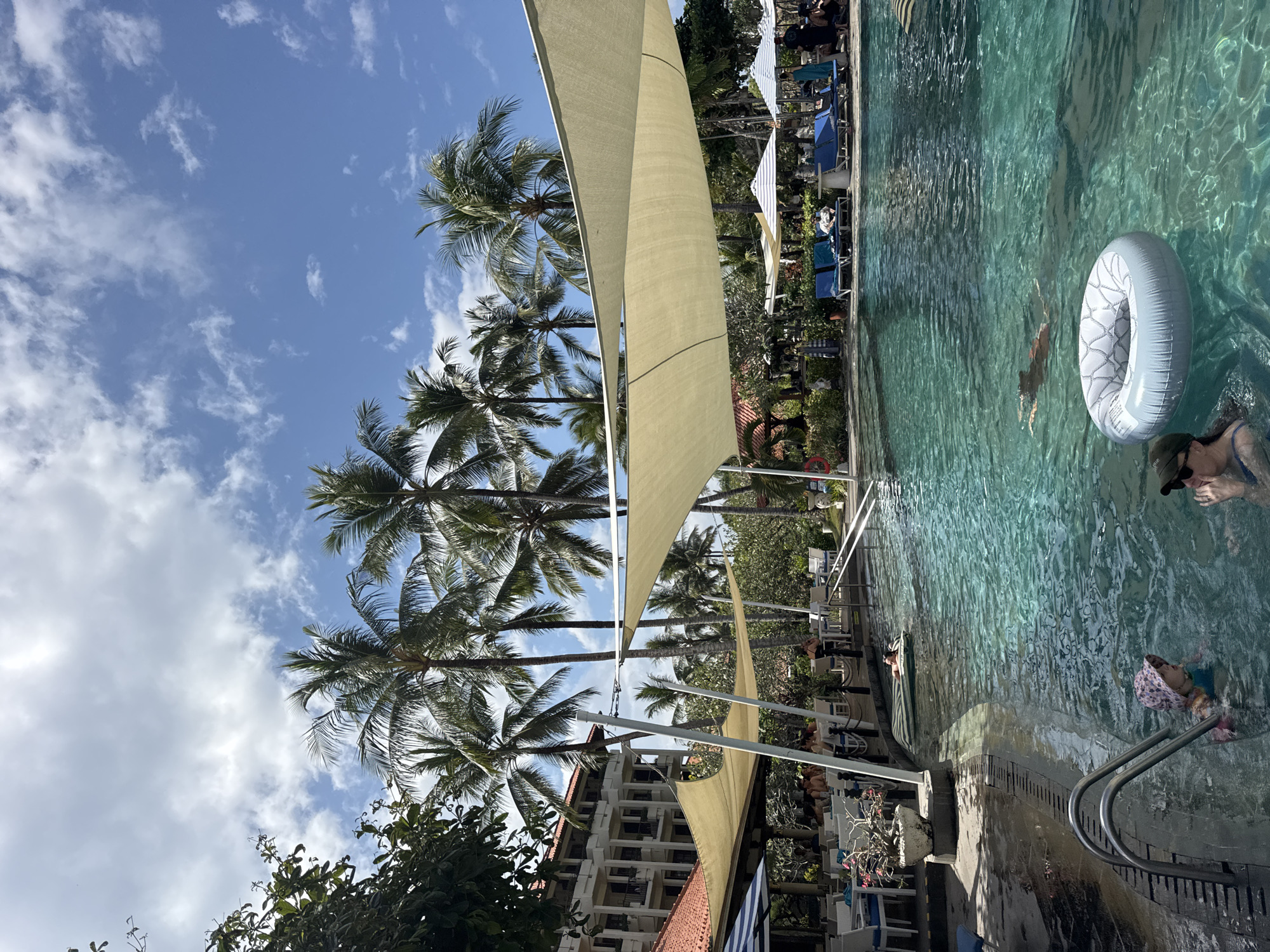 The image depicts a poolside scene at a resort in Kuta Selatan, Indonesia. A large, turquoise swimming pool dominates the foreground, with several people enjoying the water. In the lower left, a child wearing a pink hat and a blue swimsuit is partially submerged near a metal ladder. To the right of the child, an adult wearing sunglasses and a blue swimsuit is also in the water. A white inflatable ring floats in the pool, and another person is swimming further out in the water.
Shade is provided by a large, beige canopy stretched over a portion of the pool area. Beyond the pool, lounge chairs are set up, some occupied by people. Palm trees with lush fronds are visible throughout the scene, contributing to the tropical atmosphere. In the background, a multi-story building with balconies and red-tiled roofs suggests a resort or hotel structure. The sky is a clear blue with scattered white clouds. The overall environment suggests a leisure setting, likely a hotel or resort, with an emphasis on relaxation and swimming activities.