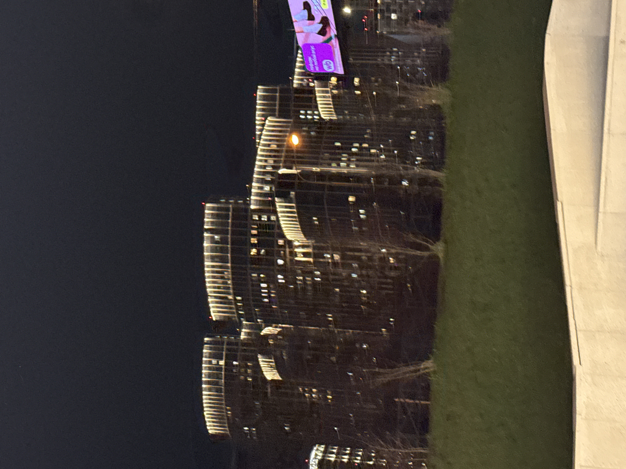 The image captures a night scene of a modern urban environment, likely in Khimki, Russia. The dominant feature is a cluster of several tall, contemporary buildings on the left, illuminated with numerous lights from within their windows and distinctive horizontal strip lighting along their facades. One building prominently displays a bright, round orange light.
Perched atop one of the buildings is a large, vibrant digital billboard. The advertisement on the billboard shows two women in what appear to be beachwear or swimsuits, set against a pink and purple background. Visible text on the billboard includes the letters "wb" in a purple rectangle, and other blurred Russian text that, upon closer inspection from the crops, reads "100% оригинал" (100% original) and "доставка бесплатно" (free delivery), strongly suggesting an advertisement for the Russian e-commerce company Wildberries.
In the middle ground, separating the buildings from a structure on the right, is a wide expanse of green grass or a lawn. Faint outlines of bare trees or branches can be seen, suggesting either winter or deciduous trees without leaves. To the far right, a large, light-colored, angular concrete or stone structure dominates the edge of the frame, possibly part of a building or a large architectural element. The sky is dark, confirming the nighttime setting. The scene is static, capturing the illuminated structures and open spaces of a developed area after dark.