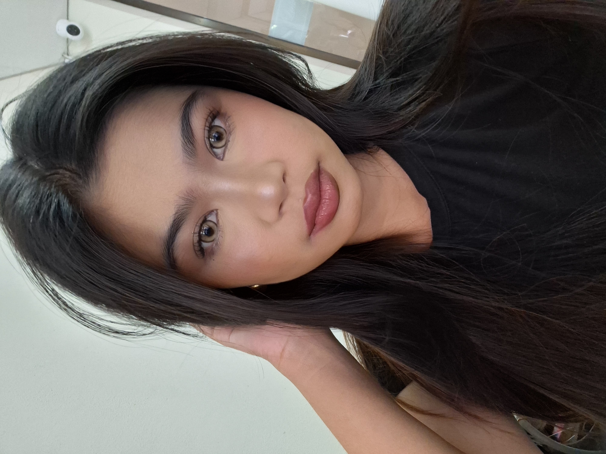 A medium close-up selfie features a young woman with long, dark brown hair styled loosely around her face and shoulders. She is wearing a black t-shirt. Her left hand is raised, touching her hair near her temple. She has light brown eyes, accentuated by subtle makeup including mascara and eyeshadow. Her lips are painted a glossy, muted berry shade. The background appears to be an indoor setting with a plain, light-colored wall to the left and a doorway or partition with some reflected light to the right. A small white security camera is visible mounted on the wall in the upper right quadrant of the image. The lighting suggests an artificial light source. The scene is devoid of any specific landmarks to pinpoint a location in Kuta, Indonesia.