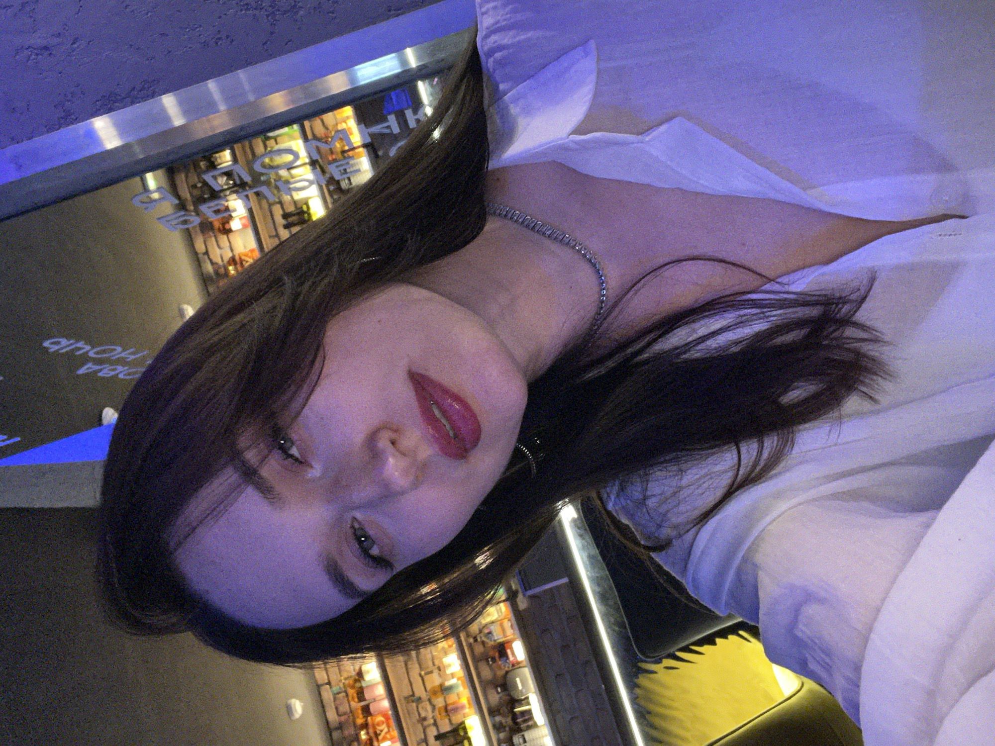 A young woman with long, dark hair, wearing a white collared shirt and a silver chain necklace, is captured in a rotated, close-up portrait, likely a selfie. Her lips are adorned with prominent red lipstick, and her gaze is directed towards the camera with a calm expression.
The setting is an indoor establishment with modern, atmospheric lighting. Cool blue and purple hues dominate the overhead and accent lighting, contrasting with warmer yellow and orange light reflecting from shelves filled with bottles or products in the background, suggesting a bar, club, or a late-night venue.
Reflected and reversed Cyrillic text is visible on a surface in the background. The clearest phrase reads "ОБА ЧАСА НОЧИ" (OBA CHASA NOCHI), which translates to "TWO O'CLOCK AT NIGHT," providing a specific time cue for the scene. The overall ambiance indicates a nighttime or late-evening social setting.