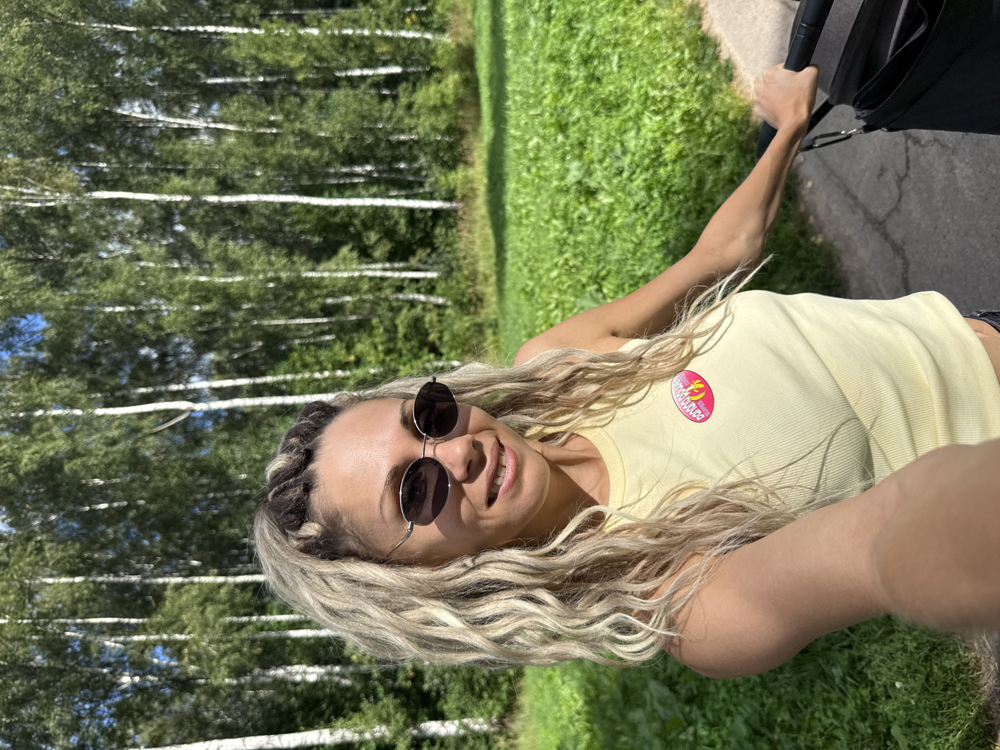 A woman with blonde dreadlocks and oval sunglasses poses for a selfie while holding the handle of a stroller. She is wearing a yellow ribbed tank top with a pink and yellow banana graphic. The background consists of a grassy field and a dense forest of birch trees under a blue sky. The woman is standing on a paved path. The scene appears to be outdoors in a park or wooded area.