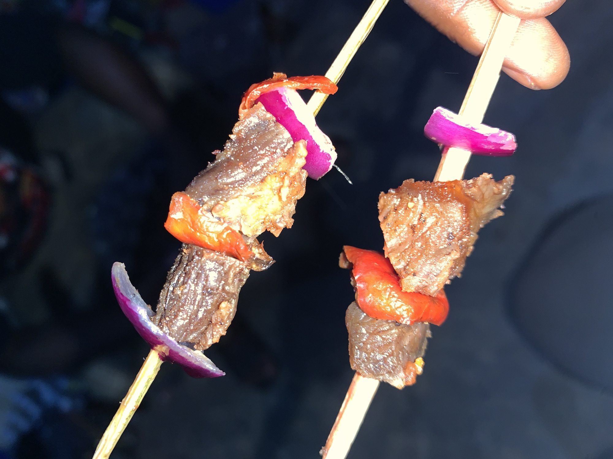 Two skewers of grilled meat and vegetables are held in a person's hand. Each skewer contains chunks of meat interspersed with pieces of red pepper and red onion. The skewers are made of light-colored wood. The background is dark and indistinct, but partially visible are other people in the background. The location is identified as New Stadium, Nigeria.