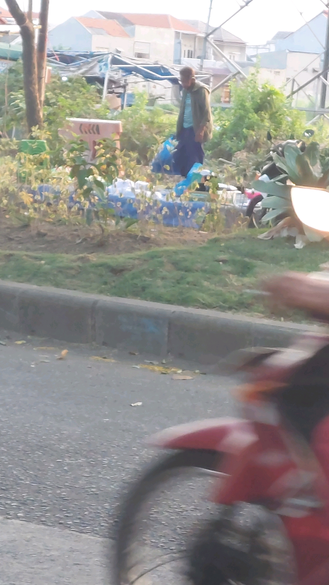 In Sepande, Indonesia, a man in a green jacket and dark pants is seen amidst scattered blue plastic sheeting and various objects. He is holding two blue plastic bags, one in each hand, and appears to be walking. The environment features overgrown vegetation, trees, and some visible structures in the background that resemble houses with orange and white roofs. A portion of a red motorcycle, including its wheel and fender, is visible in the foreground, suggesting a street or roadside location. The overall scene depicts a somewhat unkempt outdoor area with evidence of human activity and materials.