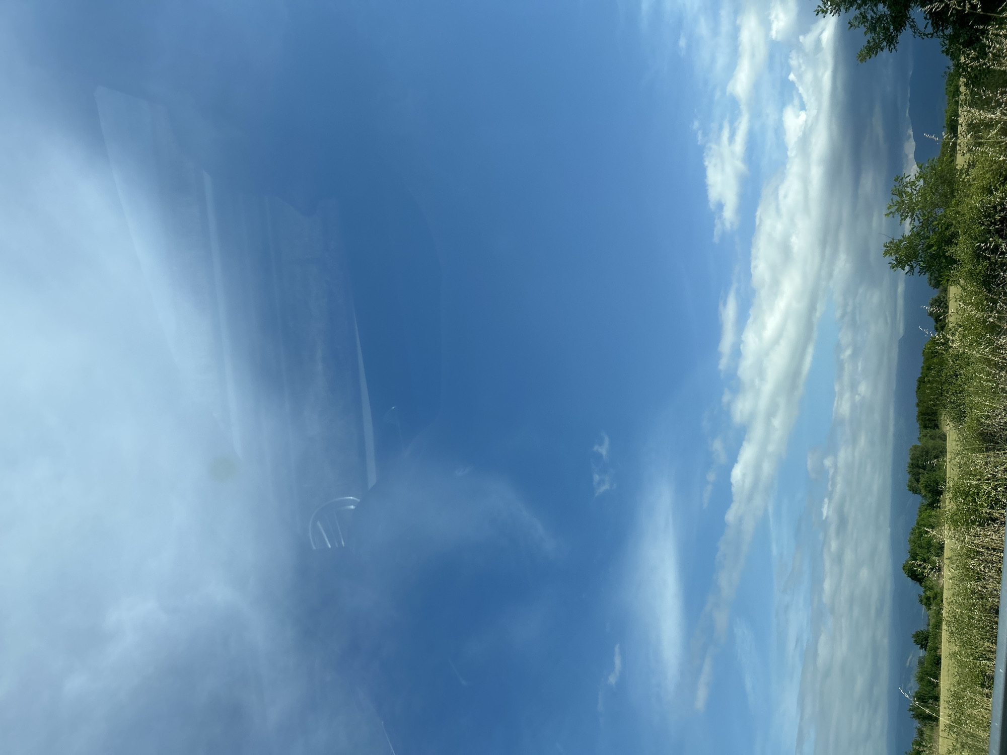 The image captures a vibrant outdoor scene, likely taken from inside a vehicle due to prominent reflections. The upper two-thirds of the frame are dominated by a clear blue sky, interspersed with scattered white, fluffy cumulus clouds and some fainter, wispy clouds.
Below the sky, a natural landscape unfolds. A line of verdant green trees and bushes stretches across the middle ground, with distant, hazy mountains visible on the horizon. To the immediate right of the landscape, a strip of tall, bright green grass or reeds occupies the foreground, suggesting a roadside view.
The left third of the image features significant reflections on what appears to be a car window. These reflections include broad areas of hazy light blue and white, mirroring the sky and clouds outside. Within these reflections, more defined, darker shapes are discernible, possibly outlining interior elements of the car, such as a headrest, or structural details reflected from the exterior.
The scene depicts a bright, clear daytime, indicating pleasant weather conditions. The strong sunlight illuminates the clouds and landscape. No people or visible text are present. The overall impression is one of serene travel through a natural, open environment, consistent with the location context of Malo Konare, Bulgaria.