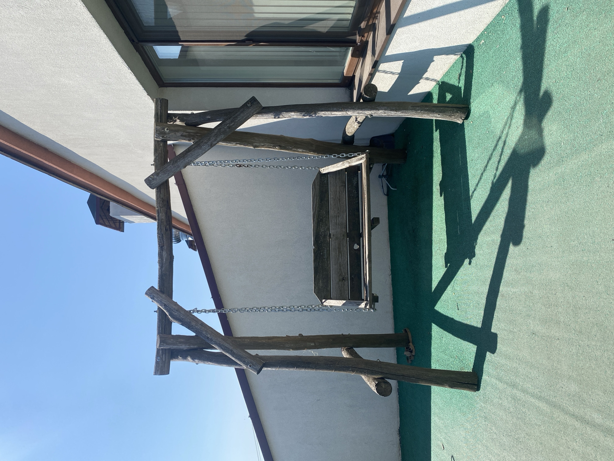 The image shows a rustic wooden swing set on a green, textured surface. The swing set is constructed from rough-hewn logs, with a wooden bench suspended by chains. The background consists of a white building with a dark-framed window and a portion of a roofline with a brown gutter. The sky is clear and blue. The scene appears to be outdoors, possibly a patio or rooftop area. There are no people or animals visible in the image. The lighting suggests it is daytime, with strong shadows cast by the swing set and the building. There are no visible indications of the specific location in Chișinău, Moldova, beyond the general setting.
