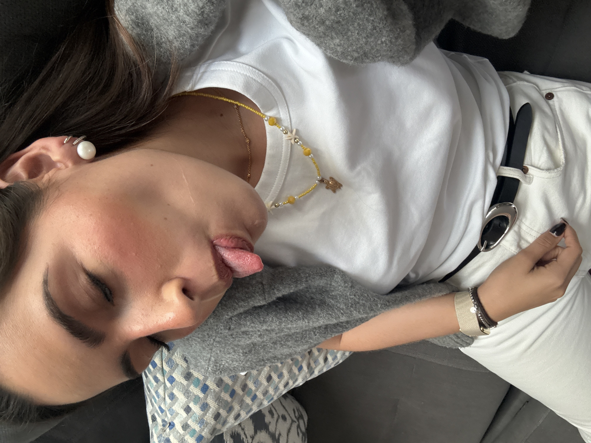 The image shows a person lying down. The person has brown hair and is wearing a white t-shirt, white pants, and a grey sweater. They are wearing a necklace with yellow and white beads, and a black belt with a silver buckle. The person's eyes are closed and their tongue is sticking out. The person is wearing several bracelets on their right wrist and an earring in their left ear. The person appears to be lying on a gray sofa with a patterned pillow. The location cannot be determined from the image.