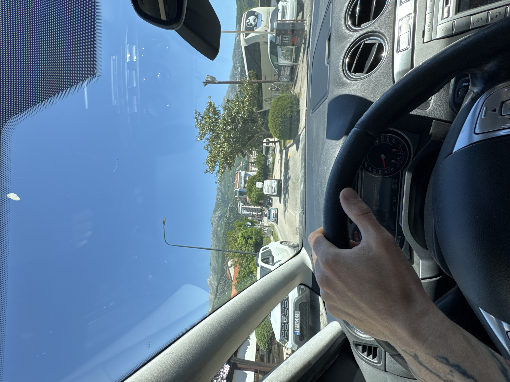 The image, taken from inside a car, shows the driver’s perspective of a street in Budva, Montenegro. The driver's left hand is holding the steering wheel. The dashboard and rearview mirror are visible, providing context for the shot. Outside, the sky is clear and bright. On the street, there are several cars, a large double-decker bus, a street light, and a parking sign. Buildings and greenery, including trees and shrubs, line the street, with mountains visible in the background. The license plate of a white Dacia Duster in front of the driver says "TV AS648" indicating that it is registered in Montenegro.