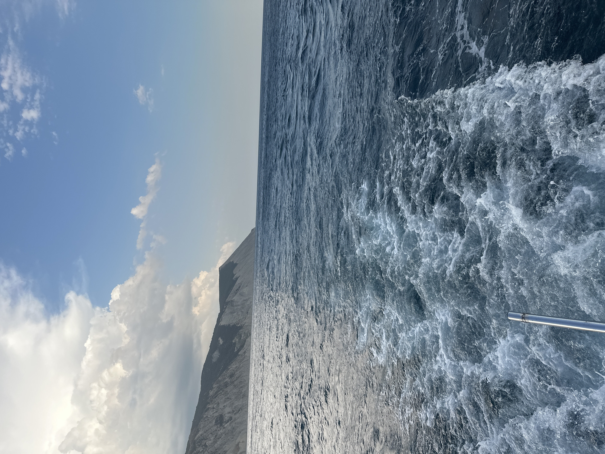 The image captures a view from a boat moving through the Black Sea, approaching the coast of Anapa, Russia. The water behind the boat is churned into a white wake, indicating movement. The sea is a deep blue, with moderate waves creating ripples and whitecaps. To the left, a rugged, treeless cliff face rises from the sea, characteristic of the Anapa coastline. The sky above is a clear blue with scattered white clouds, some of which are large and puffy, particularly over the land. A metal railing from the boat is visible in the lower foreground. The overall scene depicts a sunny day at sea with a view of the coastal landscape.