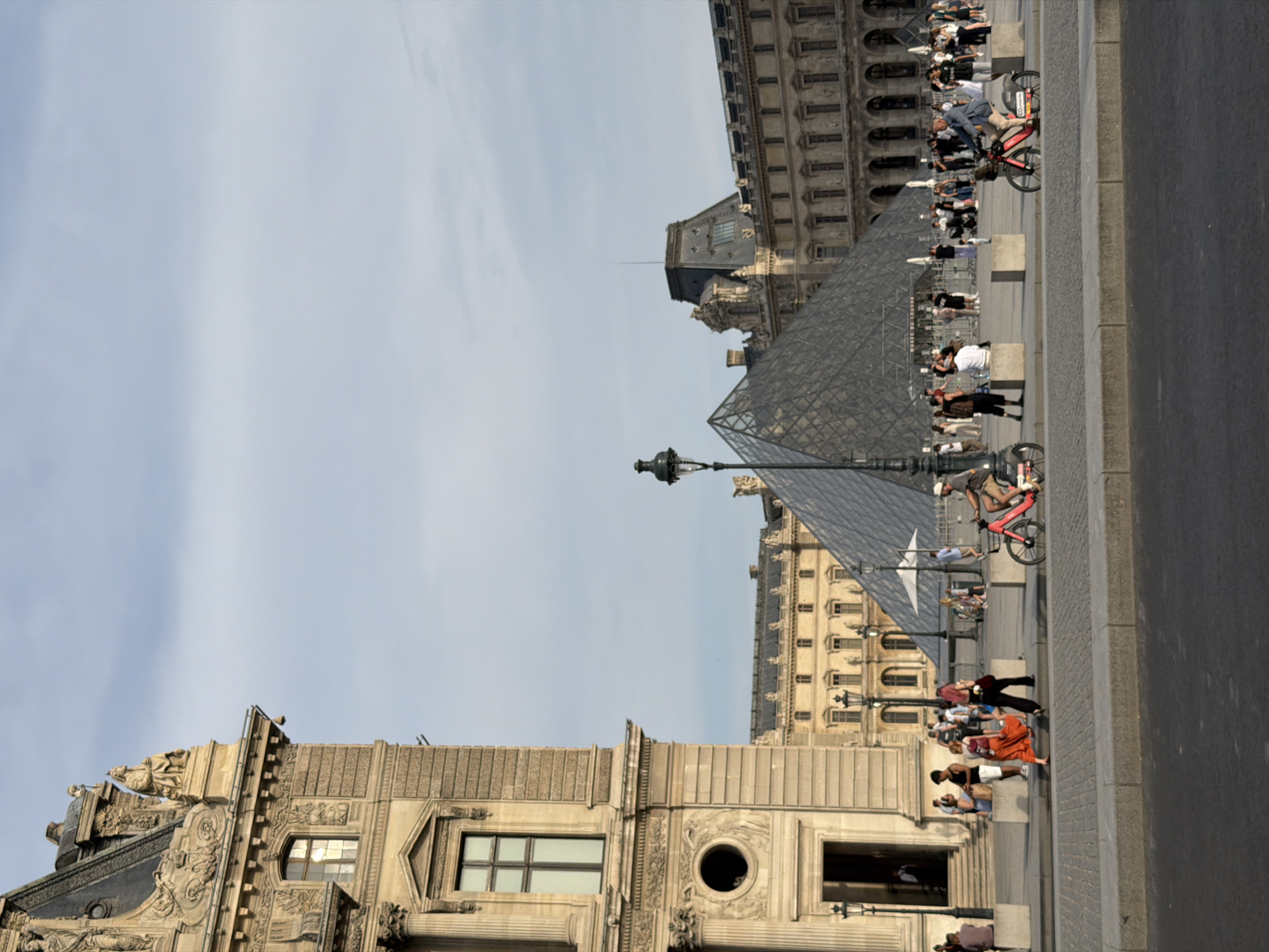 The image shows a view of the Louvre Museum in Paris, France, with its iconic glass pyramid in the background. On the left, a portion of the historic museum building is visible, featuring classical architecture with ornate detailing and large windows. In the foreground, people are walking and riding bicycles on a paved plaza. The Louvre Pyramid, a modern structure made of glass and metal, stands prominently in the center, reflecting the sky and surrounding buildings. Further back, more of the Louvre's classical facade stretches across the background, characterized by rows of windows and decorative elements. The sky is partly cloudy, with a muted blue hue. A lamppost stands to the left of the pyramid, adding to the urban streetscape. The scene depicts a bustling public space, likely a popular tourist destination, with a mix of architectural styles representing different eras of French history.