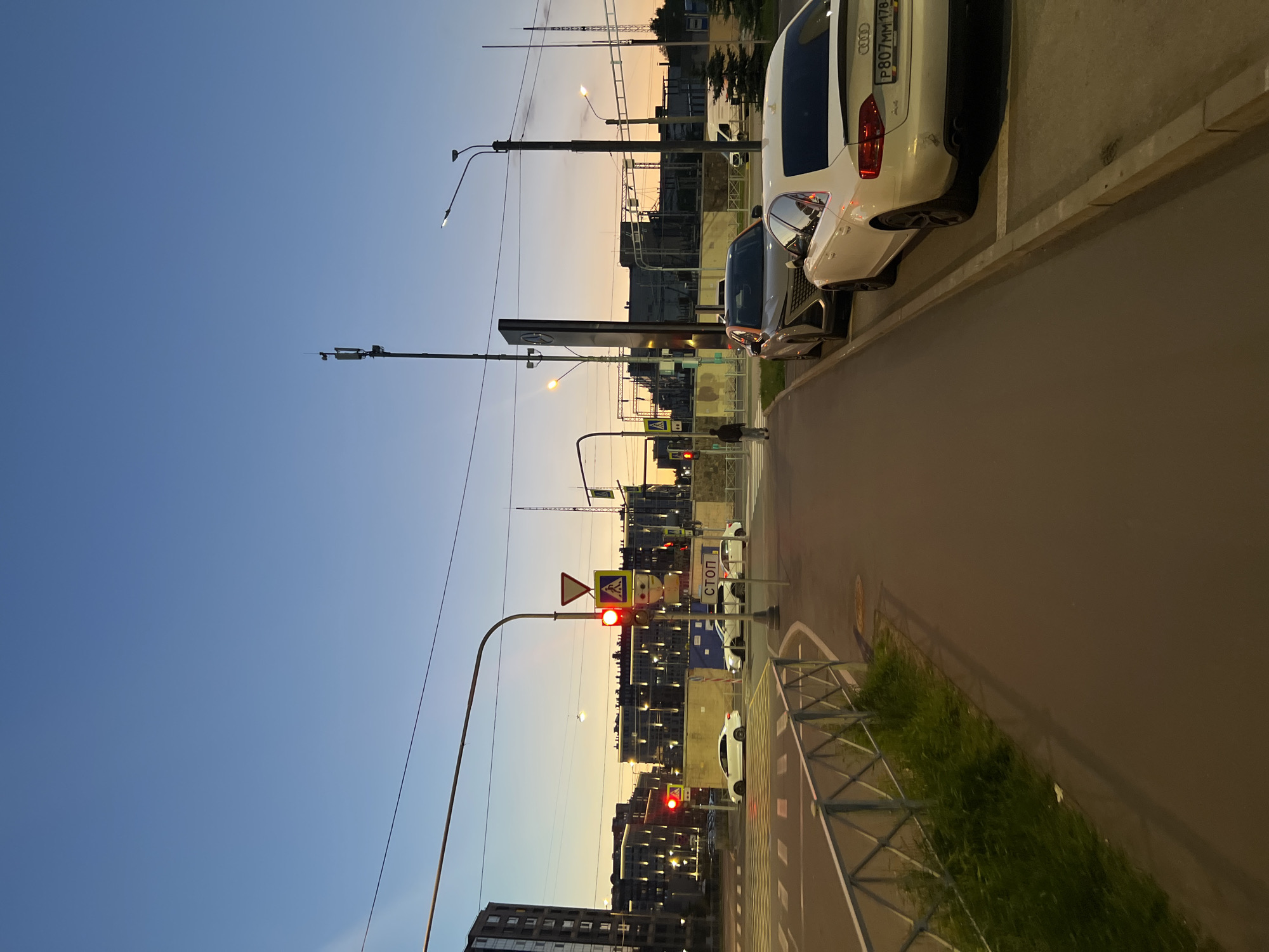 The image depicts an urban street scene at dusk in Saint Petersburg, Russia, specifically at the intersection of Komendantsky Prospekt and Ulitsa Utochkina, with the view facing southeast. The sky transitions from a deep blue overhead to warm orange and yellow hues at the horizon.
In the immediate foreground on the left, a metal pedestrian fence with a diamond pattern runs alongside a patch of tall green grass and a paved sidewalk. On the right, two vehicles are parked on a paved strip adjacent to the sidewalk. The prominent vehicle is a white Audi A4 sedan, displaying Russian license plate P807MM 178 (confirming Saint Petersburg region 178) and exhibiting minor scuffing on its rear left bumper. Behind it, a beige/gold SUV, possibly a Nissan Qashqai, is partially visible.
The midground features a wide asphalt road with clearly marked pedestrian crossings leading to an intersection. Traffic lights are illuminated red, and several road signs are visible, including a prominent red "СТОП" (STOP) sign in Cyrillic, a yellow pedestrian crossing sign, and a red yield sign. A person in dark clothing is walking away from the camera on the far side of the intersection. On the right side of the midground, a tall, dark vertical sign bearing the Mercedes-Benz logo is positioned next to a modern streetlight pole. Further down the street, a small white cargo van is partially visible.
The background is dominated by multi-story modern residential buildings on both the left and right, characterized by numerous illuminated windows. Beyond these, industrial-looking structures or power infrastructure are discernible against the twilight sky. Overhead, utility lines traverse the frame, and a cellular tower structure is visible on the right. Artificial light sources from streetlights and building interiors contribute to the illumination of the scene.