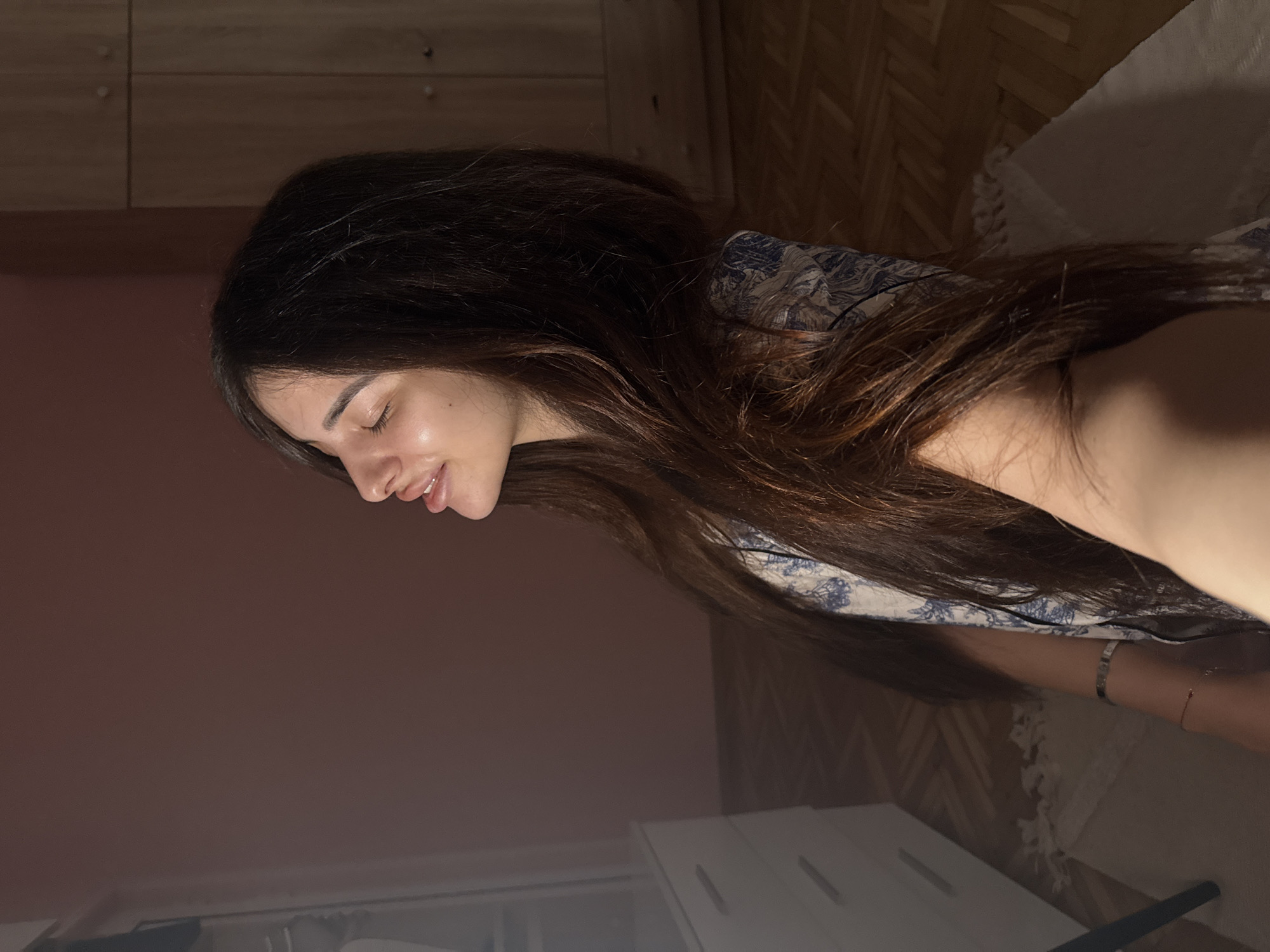 The image shows a young woman with long, dark hair, positioned in a profile view with her eyes closed and head tilted back slightly, bathed in warm light. She is wearing a patterned top. Her right arm is visible, adorned with bracelets. The background appears to be an indoor residential setting, possibly a bedroom. To the left, a white dresser or cabinet is partially visible. The flooring is wooden, with a rug underneath. In the background on the right, a wooden wardrobe with closed doors is present. The walls are painted a soft pinkish-brown. The overall atmosphere is intimate and relaxed. There is no information about the location being in Plovdiv, Bulgaria within the image or the provided crops.