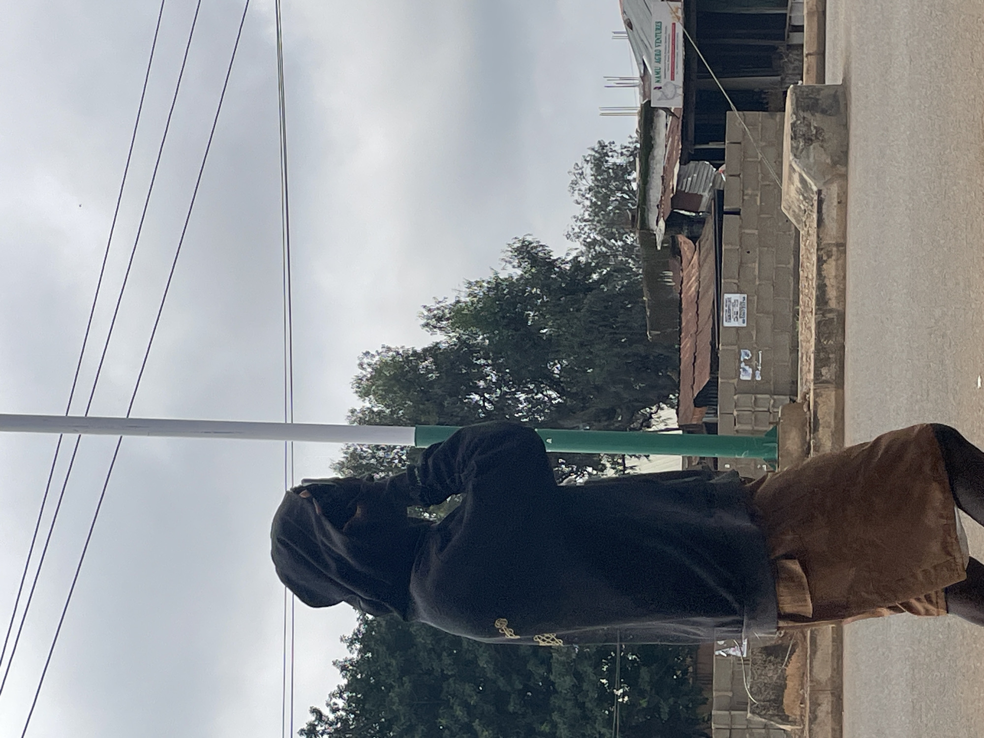 A person wearing a dark hooded sweatshirt and brown shorts stands on a paved road, looking away from the camera with their hands raised to their face, possibly shielding their eyes. The person is positioned near a green and white pole. In the background, there are trees, a low brick wall, and several buildings with corrugated metal roofs. A sign on one of the buildings reads "NAMU AGRO VENTURES". The sky is overcast. The location is identified as Bukuru, Nigeria.