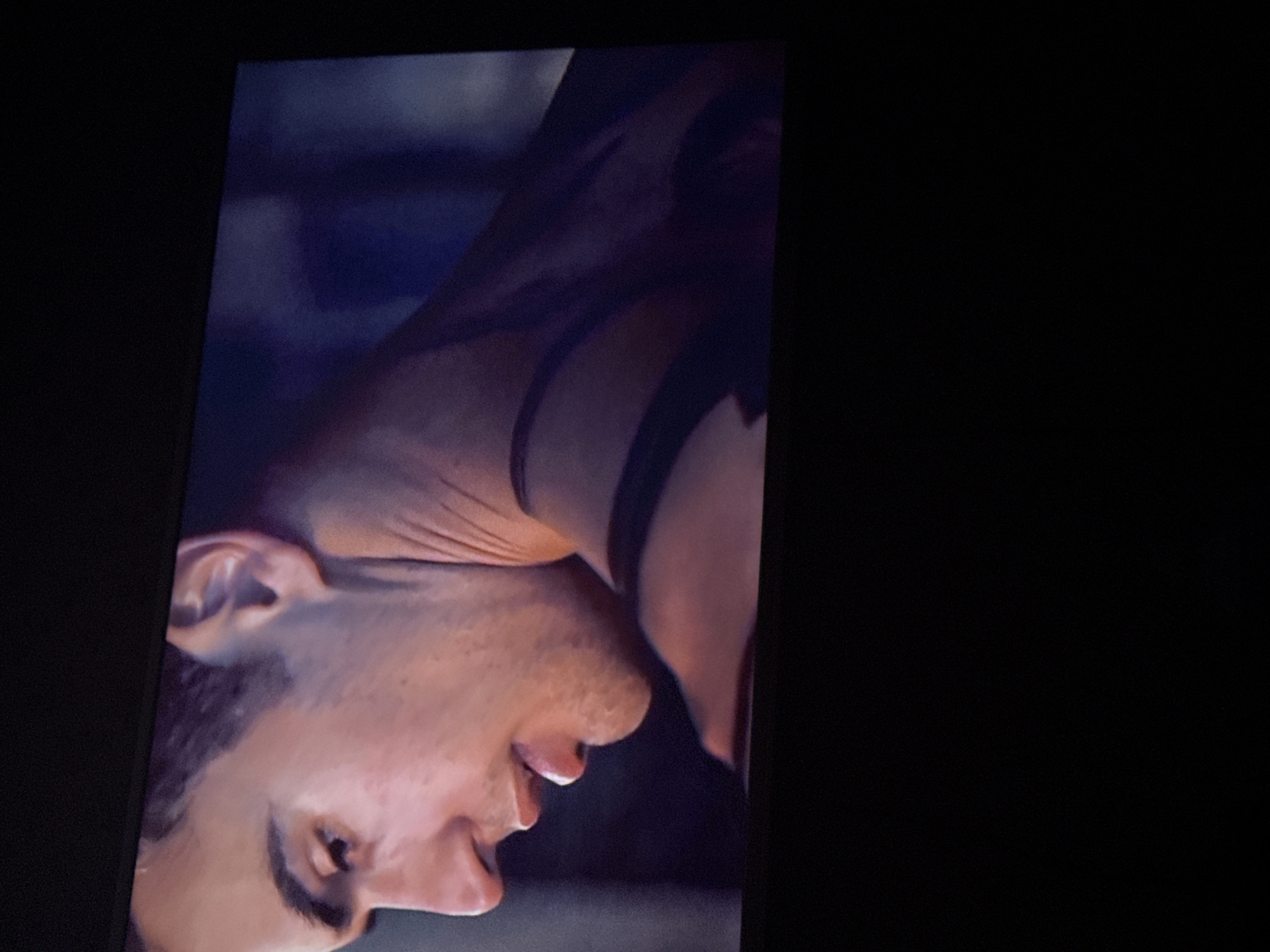 A close-up shot depicts the face of a light-skinned male with dark hair. His head is angled downward, and the camera captures his ear, nose, mouth, and part of his neck. There appear to be markings or tattoos on his neck. The background is blurred and indistinct. There is no information available to establish the location as Mytishchi, Russia.
