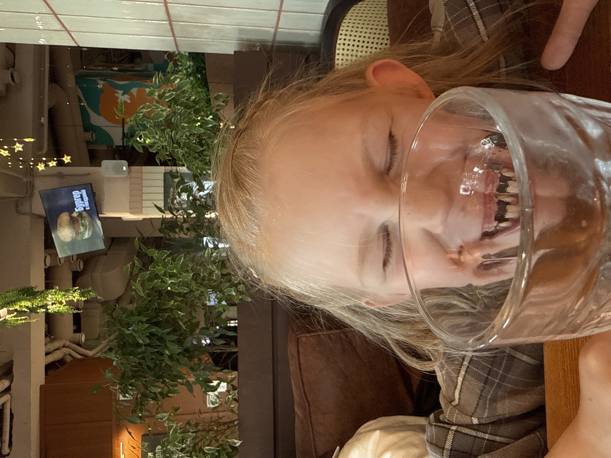 The photo, rotated 90 degrees counter-clockwise, captures a close-up of a young person's face through a clear glass. The person, with blonde hair and fair skin, has their eyes closed, and their mouth and teeth are distorted and magnified by the glass, creating a playful or comical effect. They appear to be drinking from or looking into the glass. A plaid shirt or jacket is visible on their upper body.
In the background, elements suggest an indoor setting, likely a restaurant or cafe. To the left, star-shaped fairy lights are strung across a dark area, indicating dim or evening lighting. Green foliage, possibly artificial plants, adds decor to the space. Further back, a sign with Cyrillic text is partially visible, reading "Бургер с..." (Burger with...) which appears to be a menu item. Exposed pipes or ducts and various surfaces with textures like brick or wood are also present, contributing to an industrial-chic or casual dining atmosphere. The person's left ear is visible, and behind them, part of a woven-back chair or booth can be seen. The overall impression is one of a candid moment captured in a relaxed environment.