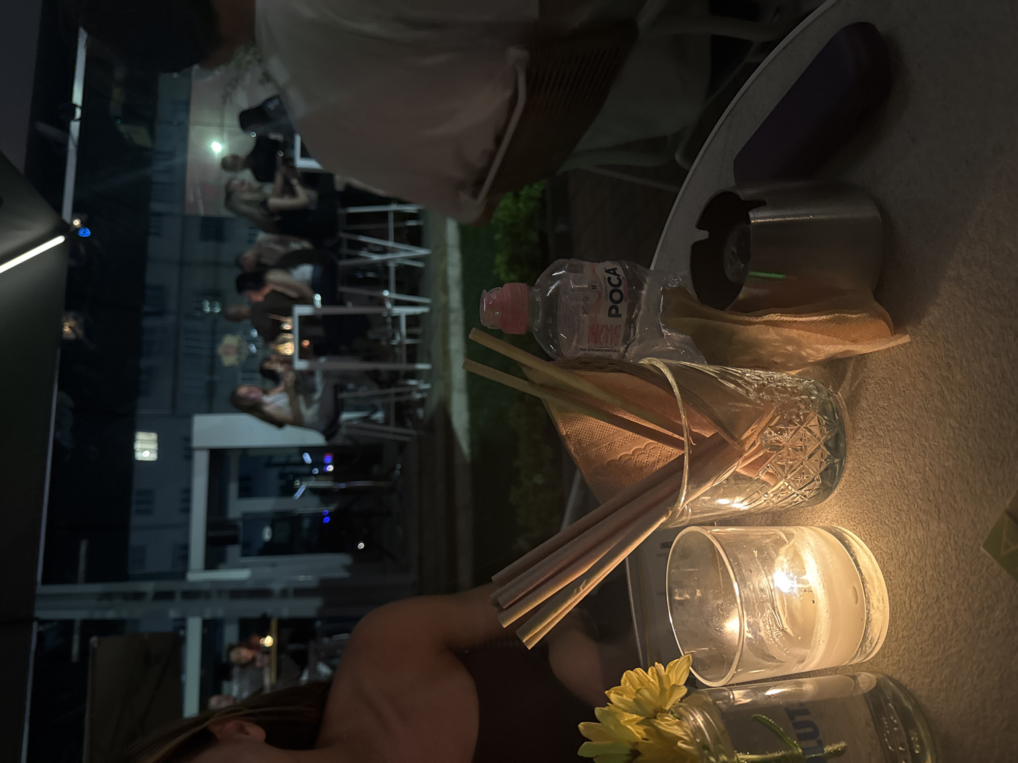 A medium shot captures a table at an outdoor restaurant at night, likely in Sofia, Bulgaria, with patrons in the blurred background. In the foreground, a lit candle flickers in a glass holder, casting a warm glow on a textured tablecloth. Next to it, a glass jar holds a single yellow flower. A second glass, decorated with a diamond pattern, contains several thin, light-colored straws and a folded napkin. A clear plastic bottle of "POCA" water sits nearby. To the right, a silver ashtray rests on the table, accompanied by a dark purple object, possibly a phone case. The background shows several groups of people seated at tables, illuminated by ambient lighting, and hints at the urban environment with structures visible in the distance.