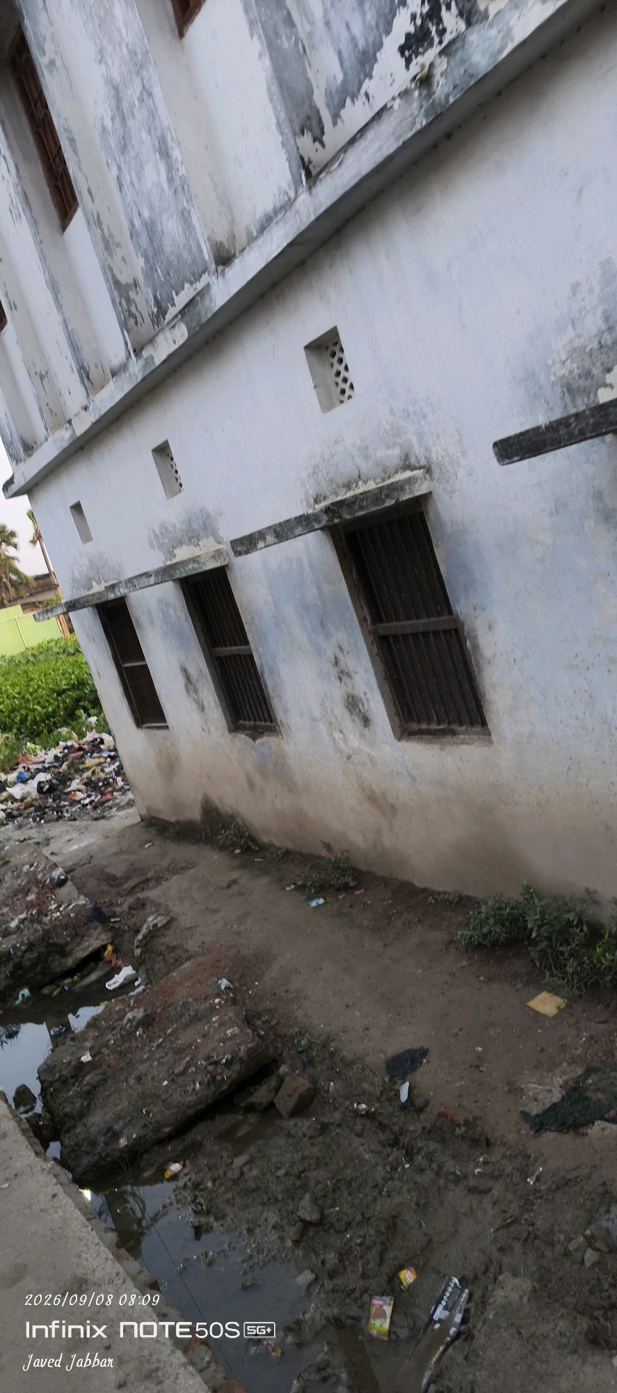 The image depicts an exterior view of a dilapidated, weathered building with peeling paint, featuring multiple barred windows and small ventilation openings. The building's wall is stained and worn, suggesting age and neglect. In the foreground, a muddy, uneven area is visible, with visible debris including plastic waste, a shoe, and other refuse scattered on the ground and in a shallow pool of water. The surrounding environment includes overgrown green vegetation and a large pile of garbage to the left of the building. The lighting suggests it is daytime. The bottom left corner of the image contains a timestamp "2026/09/08 08:09" and device information "Infinix NOTE50S SG+". The location is identified as Bahera, India.
