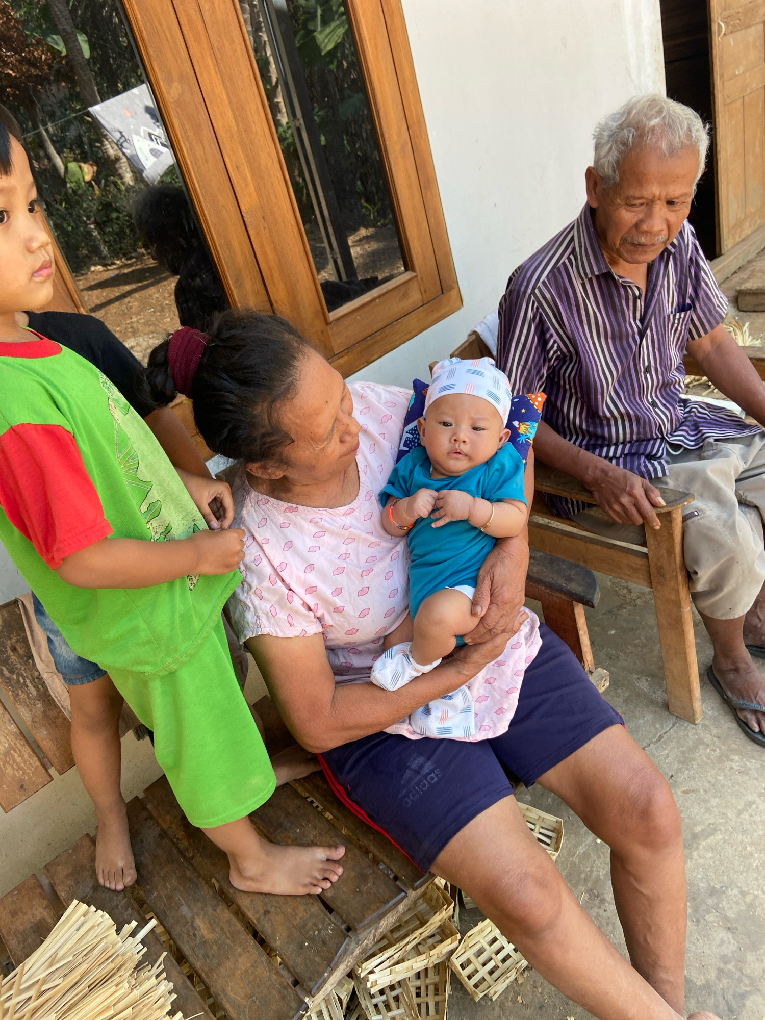In Cukilan, Indonesia, an elderly man with graying hair and a striped shirt sits on a wooden chair, looking down. Beside him, a woman holds a baby wearing a teal onesie and a patterned hat. A young child in a bright green shirt stands to the left, facing away from the camera. The scene appears to be outdoors, with wooden window frames and a white wall visible in the background. Several small woven baskets and bundles of reeds are placed on wooden slats in the foreground, suggesting a crafting or market setting.