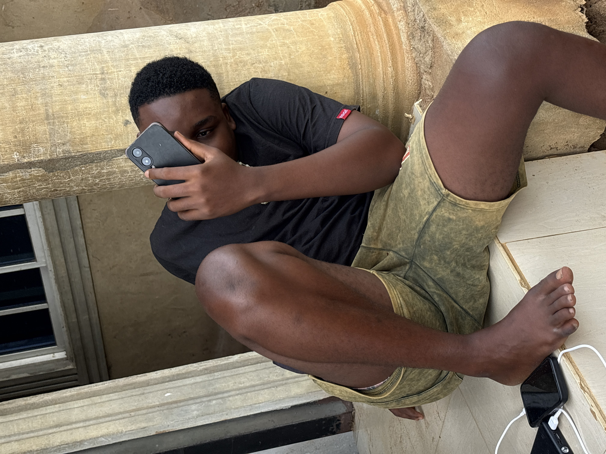 An overhead shot captures a young Black male lounging in what appears to be an indoor or semi-sheltered area in Ikot Ekpene, Nigeria. He is lying or sitting in a relaxed posture, leaning against a large, fluted, light-colored architectural column that runs horizontally across the upper and middle sections of the image.
The subject is a young man with short, dark, kinky hair and a dark skin tone. He is wearing a plain black t-shirt, which has a small red tag with white text reading "BOGE" visible on the right sleeve. He is also wearing greenish-brown, cargo-style shorts. He is barefoot, with his right foot resting on a light-colored tiled floor.
His attention is focused on a black smartphone he holds in his right hand, obscuring part of his face. The phone has a visible dual-camera setup on its rear. Another black smartphone lies on the tiled floor near his bare foot, connected by a white charging cable to a black power bank. A second white cable also emerges from the power bank.
In the background, a section of a white-framed window with dark panes is visible to the left of the man. The general lighting suggests it is daytime. The scene conveys a sense of casual relaxation.