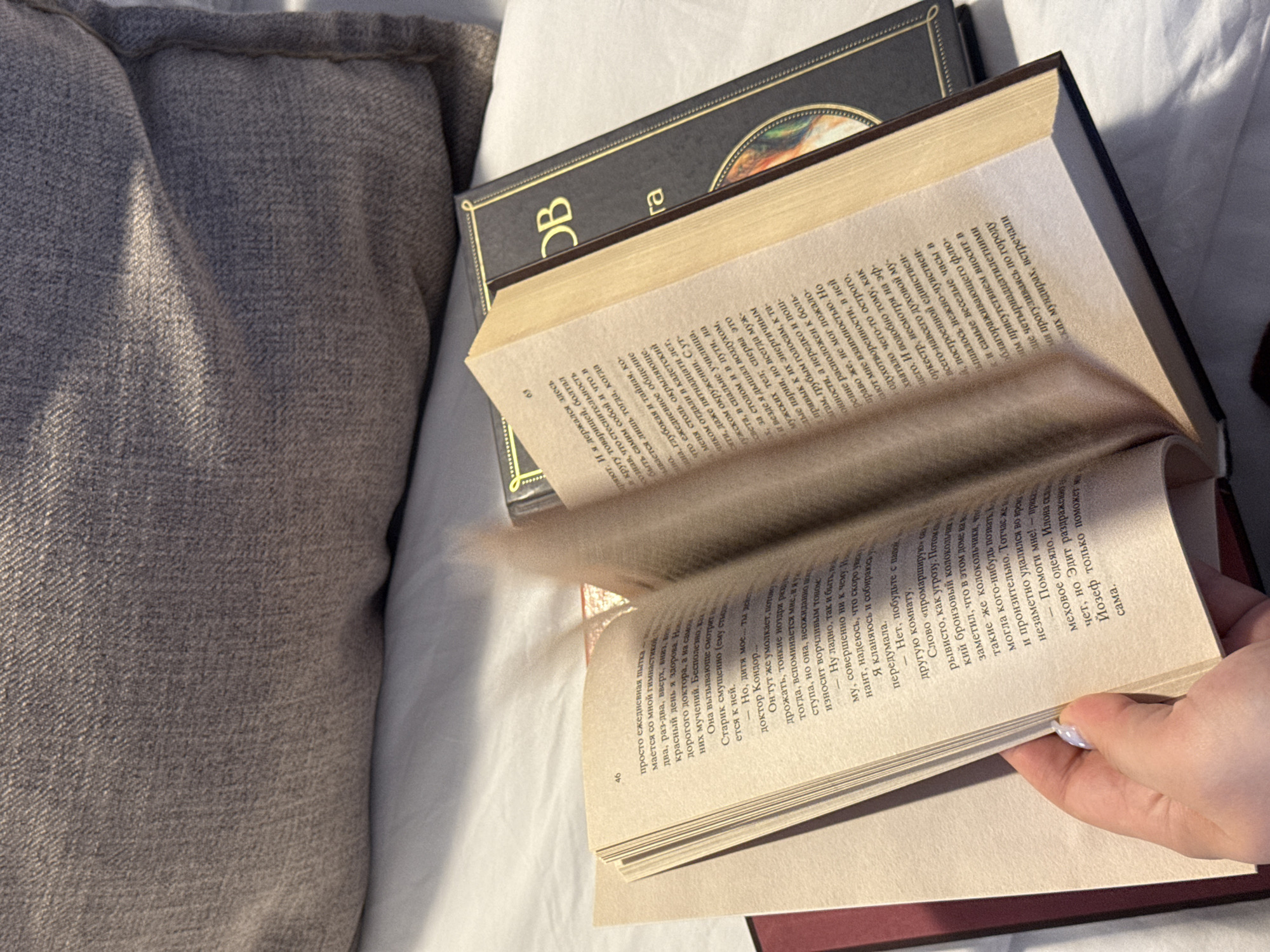 An overhead shot shows an open book being held by a hand with pale skin and a light purple, pearlescent nail polish on its thumb and index finger. The book's pages are yellowed and worn, with printed text visible. The hand is positioned at the lower left of the image, holding the book open. The book's spine is visible on the right, with a black cover and gold lettering that reads "OB" and "ra". Another book with a red binding is partially visible to the left of the open book. The scene is set on a white, textured surface, possibly a bed, with a folded, grey, textured pillow in the background. The lighting suggests a warm, possibly natural light source coming from the right. There is no visible connection to Starokorsunskaya, Russia.