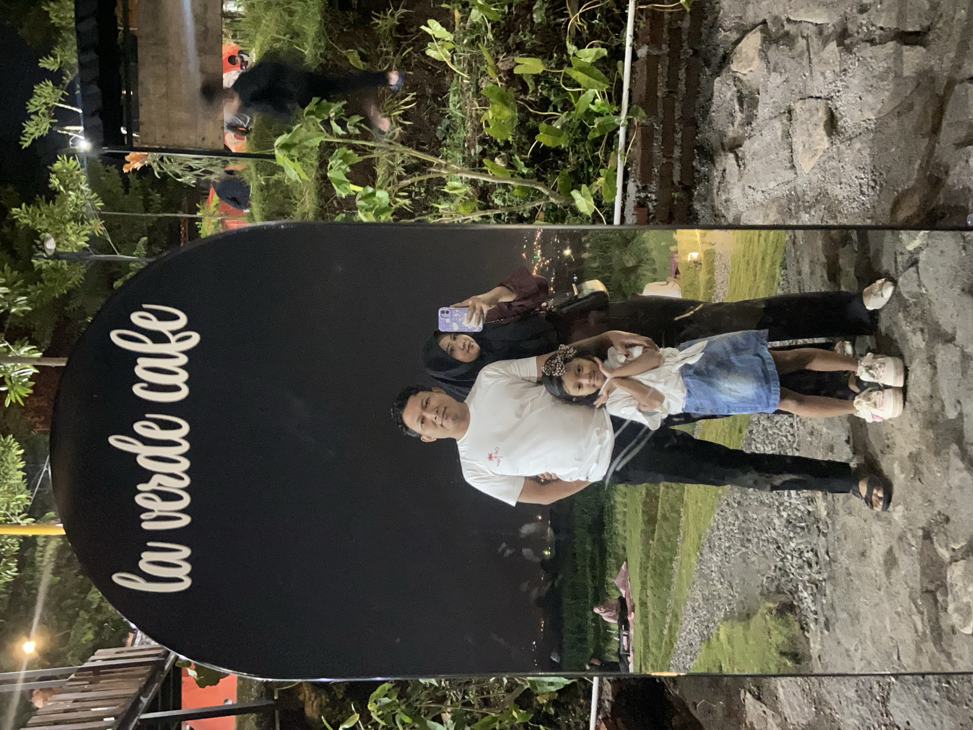 The image captures a family posing for a photograph at "La Verde Cafe" in Kesiman, Indonesia. In the foreground, a man in a white t-shirt and black pants stands next to a young girl wearing a white top and a denim skirt. The girl is positioned in front of the man, with her hands near her face. To the right of the man, a woman in a black hijab and a maroon top is taking the photo with her smartphone.
The cafe appears to be an outdoor venue, with tiered grassy areas in the background where other patrons are seated on picnic blankets. The setting is illuminated by artificial lights, creating a warm ambiance. The cafe's name, "La Verde Cafe," is prominently displayed in white cursive lettering on a large, arched black sign in the foreground. The overall scene suggests a casual and enjoyable evening outing for the family.