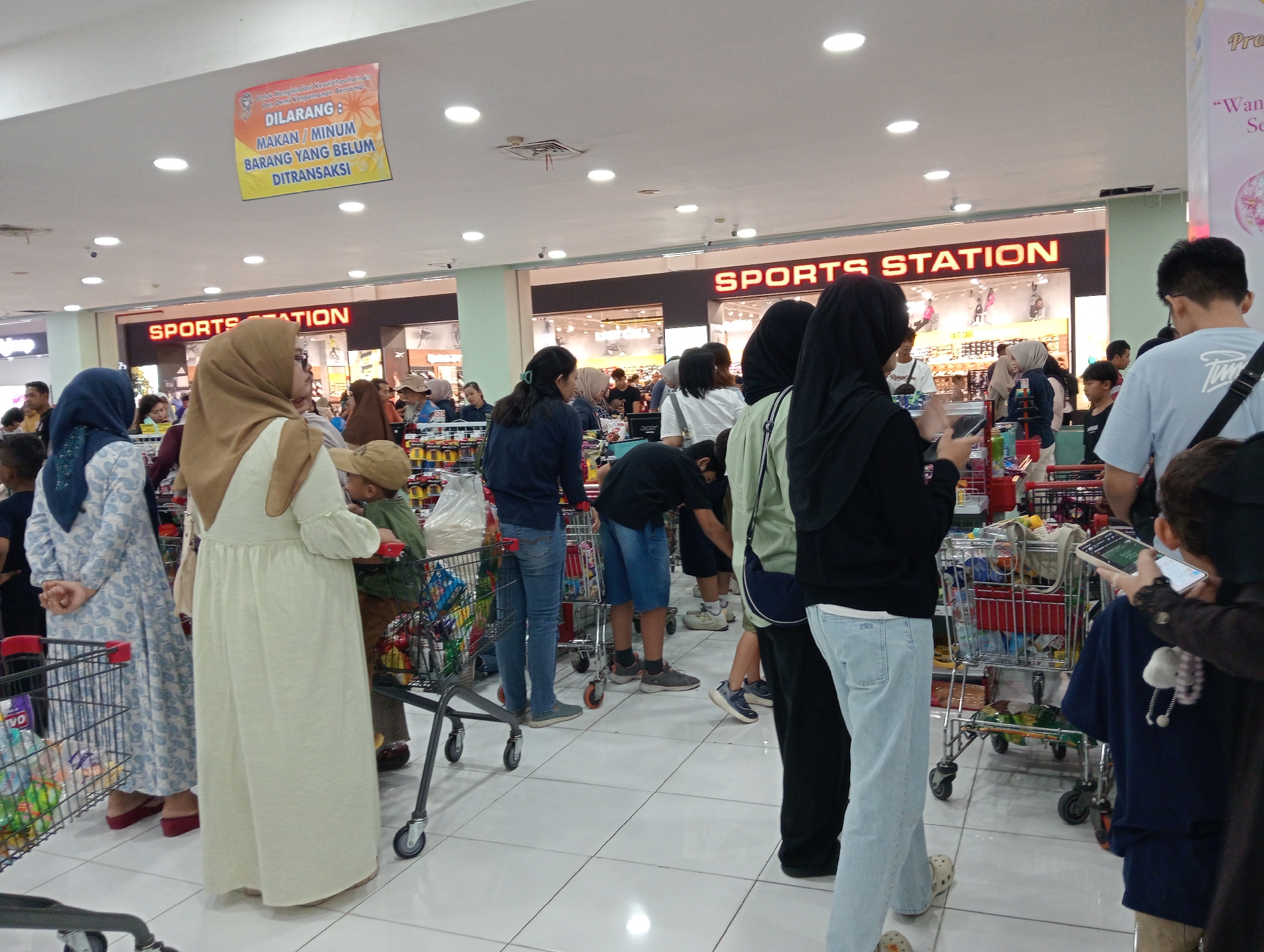 The image depicts a crowded indoor scene, likely a supermarket or department store, in Sempusari, Indonesia. In the foreground, a woman in a light-colored dress stands with her back to the camera, her left hand resting on a shopping cart. To her left, another woman in a blue and white patterned dress is also facing away from the camera. Behind them, several other individuals, including children, are visible, many wearing casual clothing. Shopping carts filled with items are scattered throughout the scene. Overhead, bright circular lights illuminate the space, and a sign in Indonesian is prominently displayed on the ceiling, stating "DILARANG: MAKAN / MINUM BARANG YANG BELUM DITRANSAKSI," which translates to "PROHIBITED: EATING / DRINKING ITEMS NOT YET TRANSACTED." In the background, the signage for "SPORTS STATION" is visible, indicating a retail establishment. The floor is tiled with white squares. The overall atmosphere suggests a busy shopping environment.