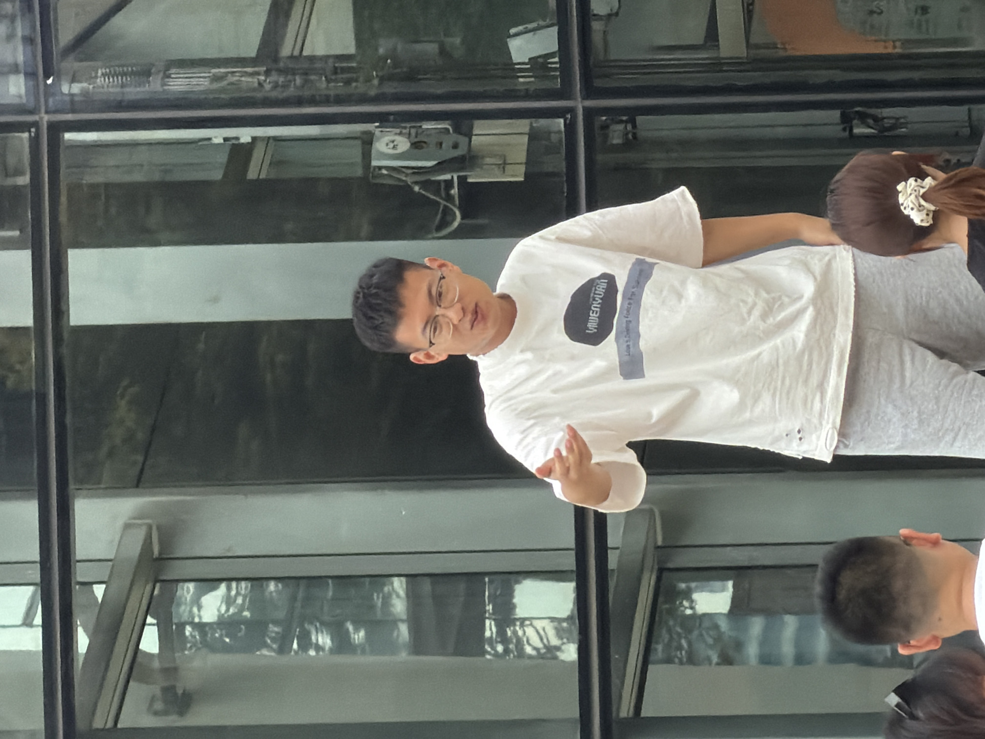 A young adult East Asian male with short black hair and glasses is standing outside, gesturing with his right hand. He is wearing a white t-shirt with a graphic print and light grey sweatpants. The background consists of a modern building with large glass windows and dark frames, possibly in Yuzhong District, China. The reflections in the glass indicate an outdoor environment. In the foreground, the backs of two individuals are visible, one with short brown hair and a patterned scrunchie, and another with short dark hair.