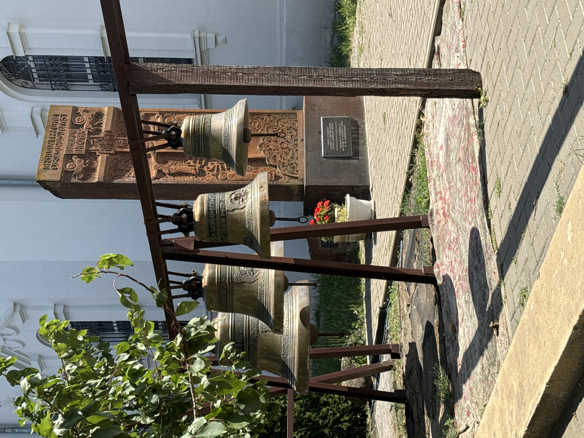 In Chaltyr', Russia, a row of four ornate brass bells hangs from a dark metal frame in front of a large, light-colored building. A stone monument with intricate carvings and inscriptions stands behind the bells. A tree with vibrant green leaves branches out from the left side of the frame, casting shadows on the ground. The area is paved with light gray bricks, and a patterned rug lies on the ground beneath the bells. A white planter with red flowers is visible to the right of the monument, adding a splash of color to the scene. The overall setting appears to be a courtyard or a public space outside a place of worship or historical significance.