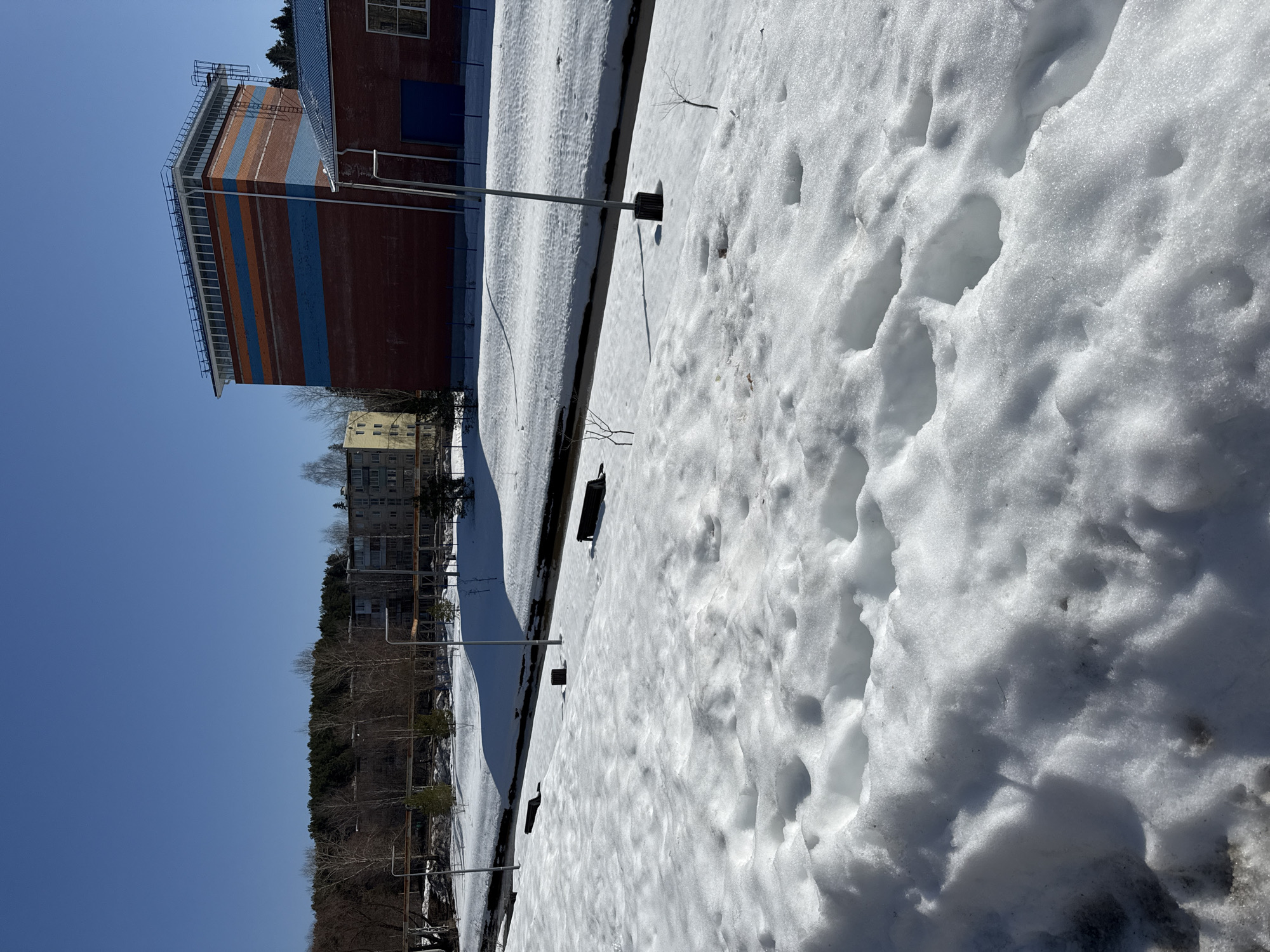 The image shows a snowy landscape in Votkinsk, Russia. Footprints are visible in the snow-covered ground. A tall, multi-story brick building with orange and blue horizontal stripes is present on the left side. Nearby is a residential building. A line of bare trees and vegetation can be seen. The sky is clear and blue.