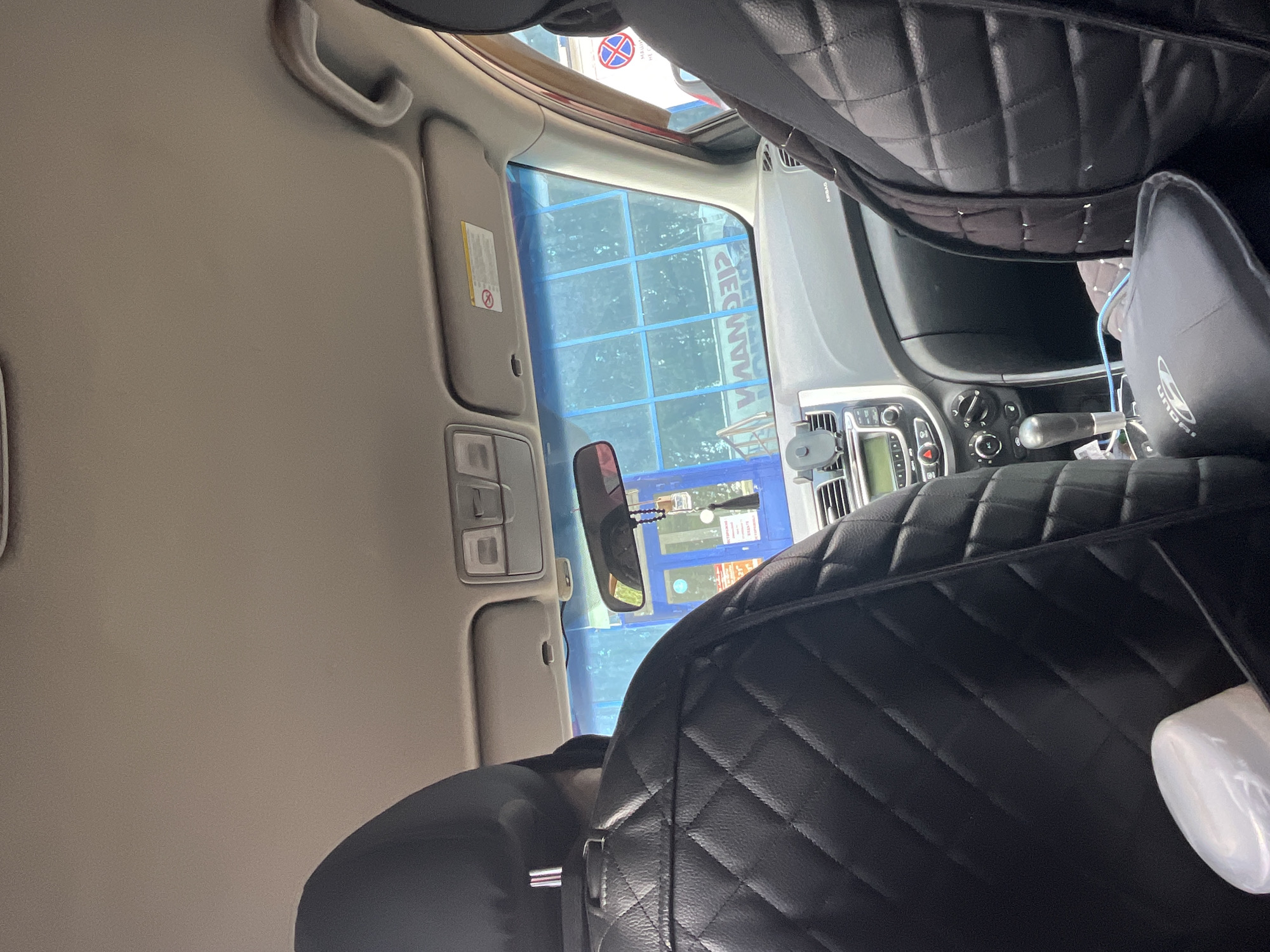 The image is taken from inside a car, looking forward through the windshield. The interior of the car features black, quilted seat covers. The dashboard is visible, with a car stereo and air conditioning controls. A phone mount is attached to the dashboard. Outside the car, a blue building with large windows is visible. There appears to be some sort of service area or establishment with signage in Cyrillic, one sign reads "СТОП" (STOP). The sky is visible through the tinted windshield. The scene is likely a street or parking lot in the city of Novaya Otradovka, Russia, possibly near a car service center.