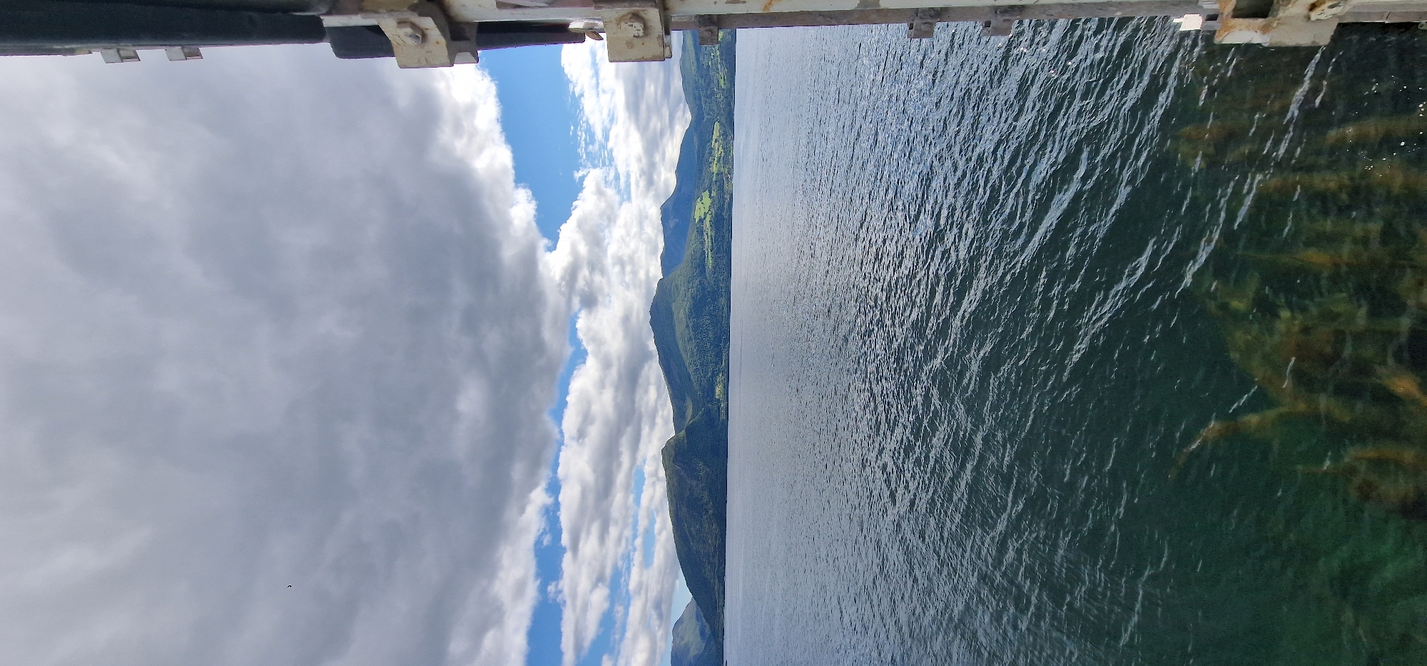 The image shows a wide shot of a body of water, likely a fjord, with a distant mountainous landscape under a partly cloudy sky. On the right side of the image, a portion of a metal structure, possibly part of a bridge or a ferry, is visible. The water has ripples and is a dark blue-green color. The distant mountains are covered in lush green vegetation, with some patches of lighter green that may indicate fields or clearings. The sky has a mix of dark grey clouds and bright blue patches, suggesting intermittent sunlight. There are no people visible in the image. The scene appears to be captured from a moving vessel or a fixed elevated point overlooking the water. The location is Volda, Norway, as indicated by the reference to the city in the prompt.
