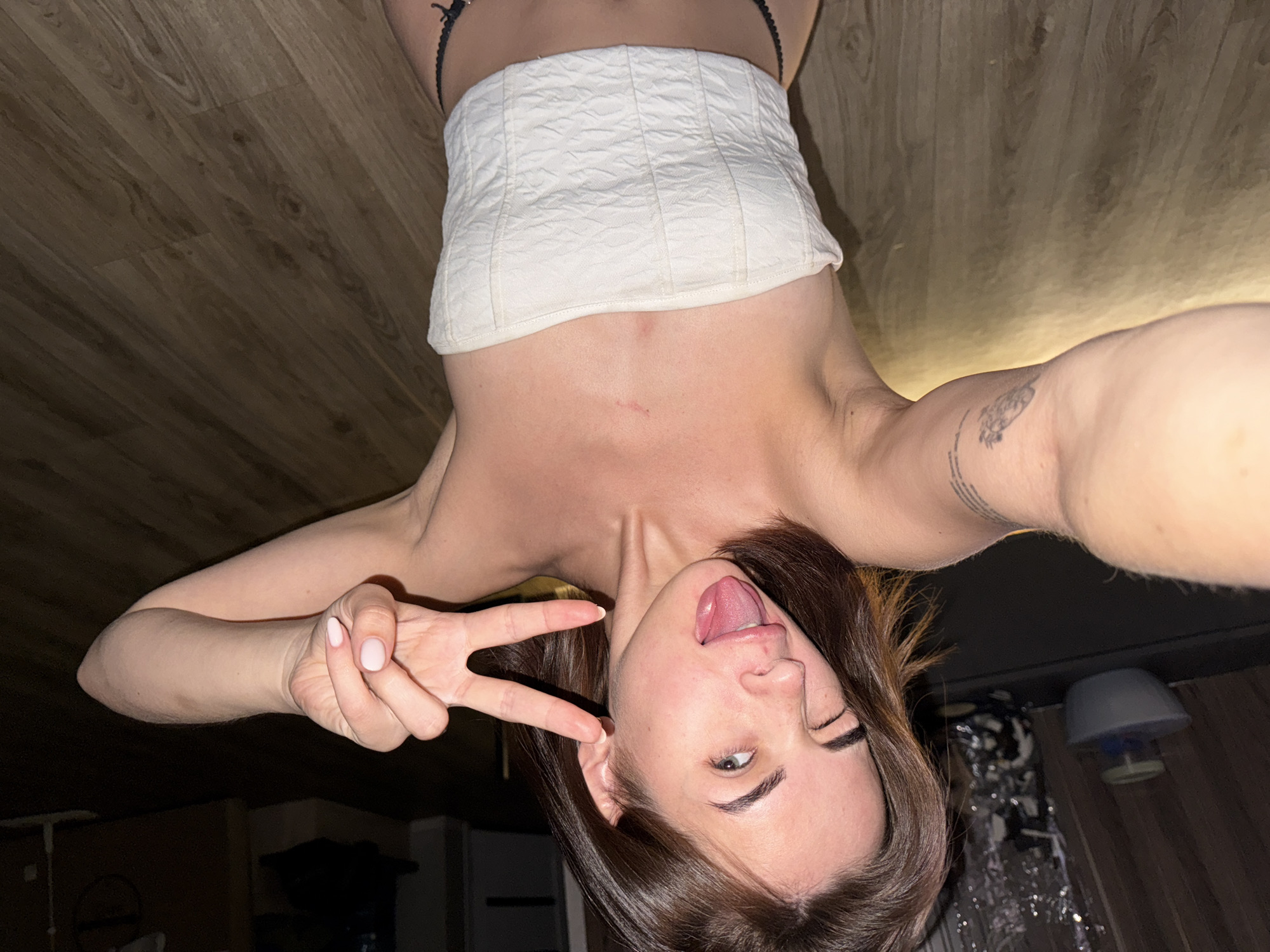 A person, presumably female, is upside-down in a selfie. She's wearing a white strapless top and black underwear, sticking her tongue out and giving a peace sign. Tattoos are visible on her right arm. She has brown hair and fair skin. The background features a wood floor and a glimpse of a room with household objects, including a lamp. The image location is not identifiable from the image.