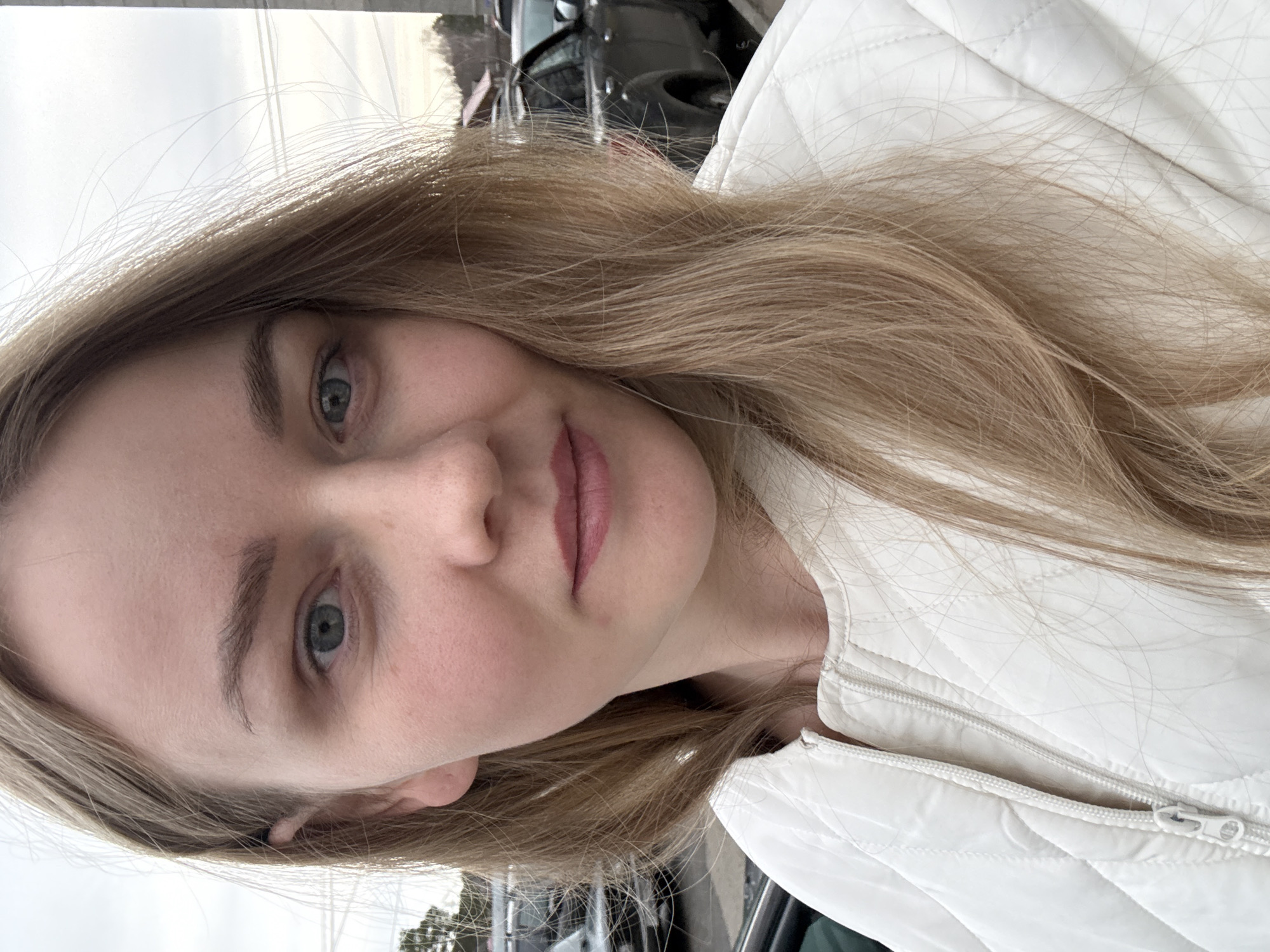 A fair-skinned woman with blonde hair and blue eyes is pictured in a close-up, tilted selfie. She is wearing a white quilted jacket with a visible zipper. The background is blurred, showing vehicles and sky, likely indicating an outdoor setting. There is insufficient information to determine whether the image was taken in Chaikovsky, Russia.