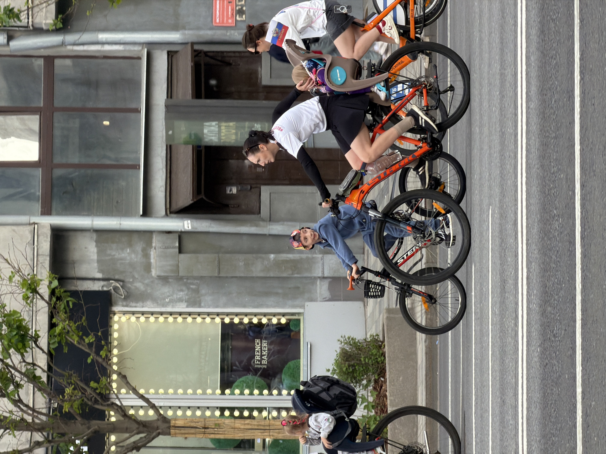 The image shows four people riding bicycles on a road in Moscow, Russia. The cyclists are riding in a line, with one adult carrying a child in a child seat, another adult, a young boy, and a toddler. The riders are wearing casual clothing and helmets. The backdrop consists of buildings with a business named "French Bakery." The road appears to have designated bicycle lanes marked with white lines. A tree is on the left edge of the image.