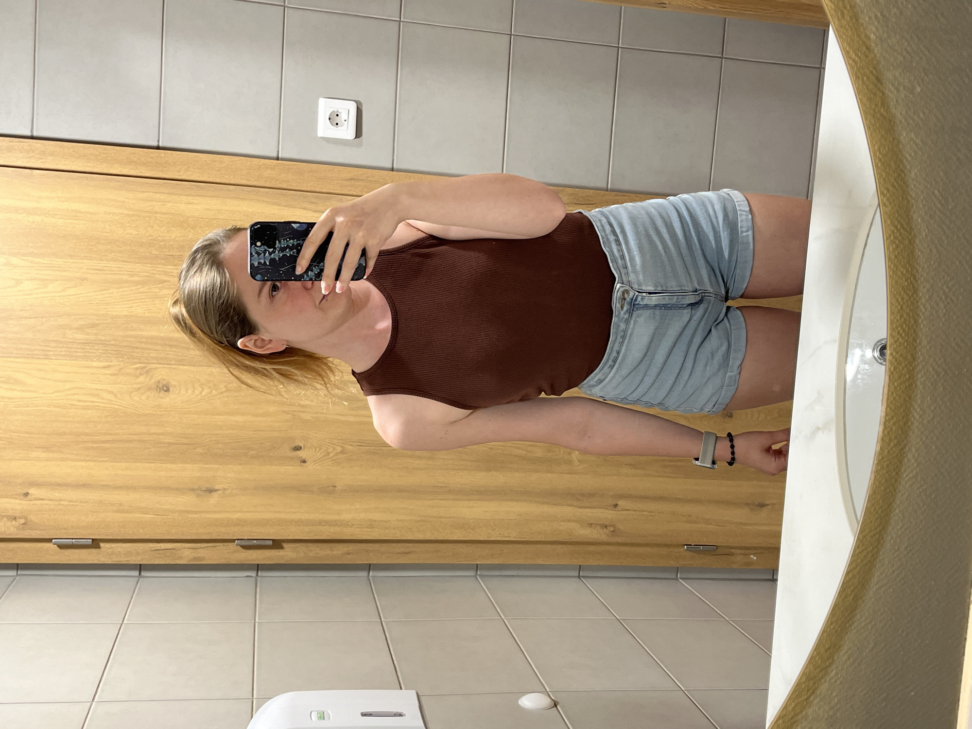 A young woman with blonde hair tied in a ponytail takes a mirror selfie in a public restroom. She is wearing a brown ribbed tank top and light blue denim shorts. She has a smartwatch on her left wrist and is holding a smartphone with a floral case. The restroom has tiled walls, a wooden door, and a white sink. The image appears to have been taken in a public restroom, possibly in a restaurant or hotel, in Plovdiv, Bulgaria.
