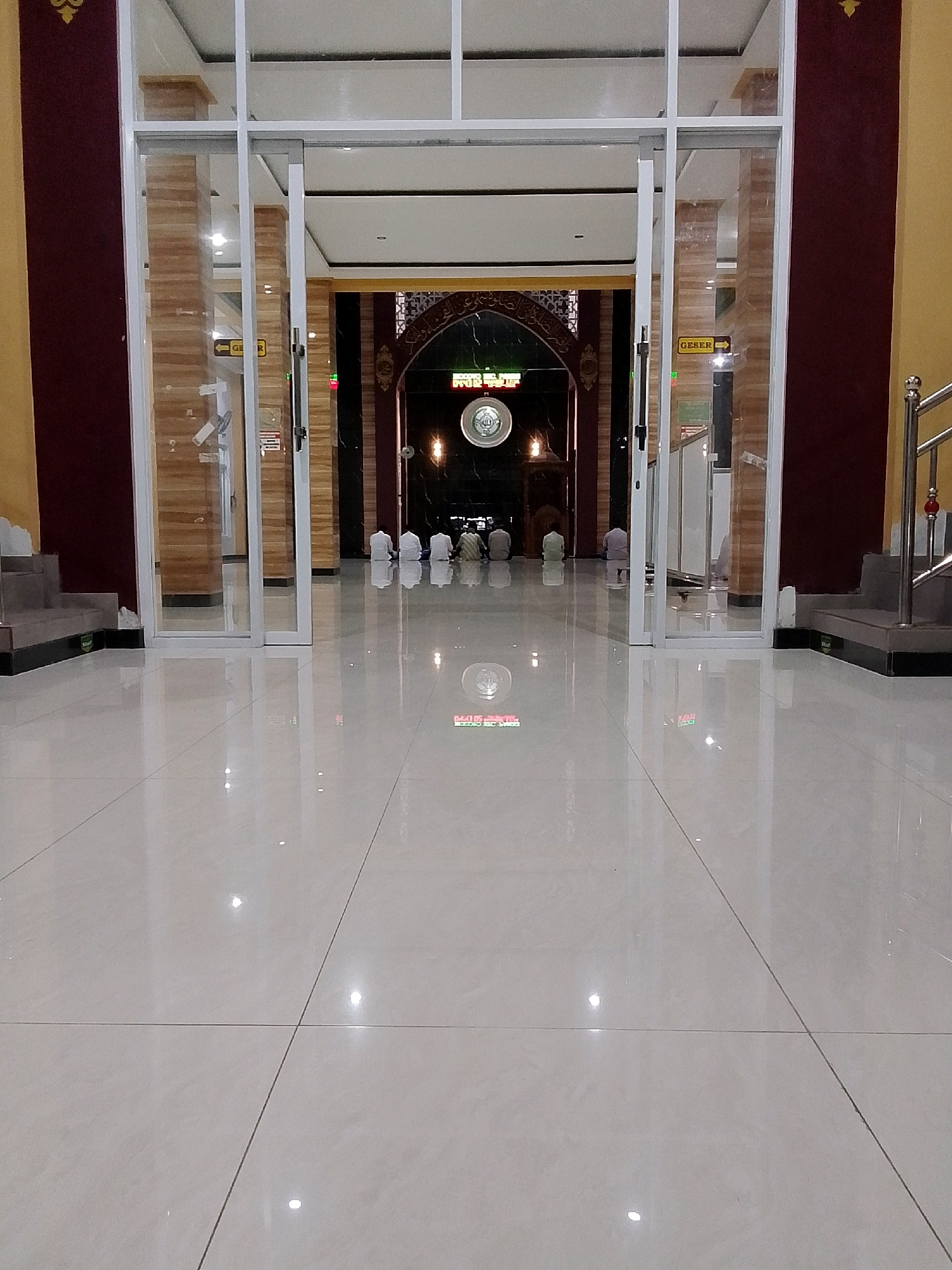 The image depicts the entrance to a mosque in Wonocoyo, Indonesia. The perspective is from the ground level, looking towards the mosque's interior through a set of glass doors. The floor is made of polished white tiles that reflect the overhead lighting, creating a glossy and bright appearance.
Inside the mosque, several people are visible, kneeling on prayer mats in prayer. They are dressed in traditional modest attire, and their backs are towards the camera. The interior of the mosque features patterned walls and decorative elements above the entrance. A circular emblem with Arabic calligraphy is prominently displayed above the doorway.
To the left of the entrance, a staircase is visible, leading upwards and away from the camera. The walls of the building are painted in a deep red color. The overall scene suggests a place of worship that is clean, well-maintained, and actively used by the community. The lighting is bright, and the reflective surfaces contribute to a sense of spaciousness.