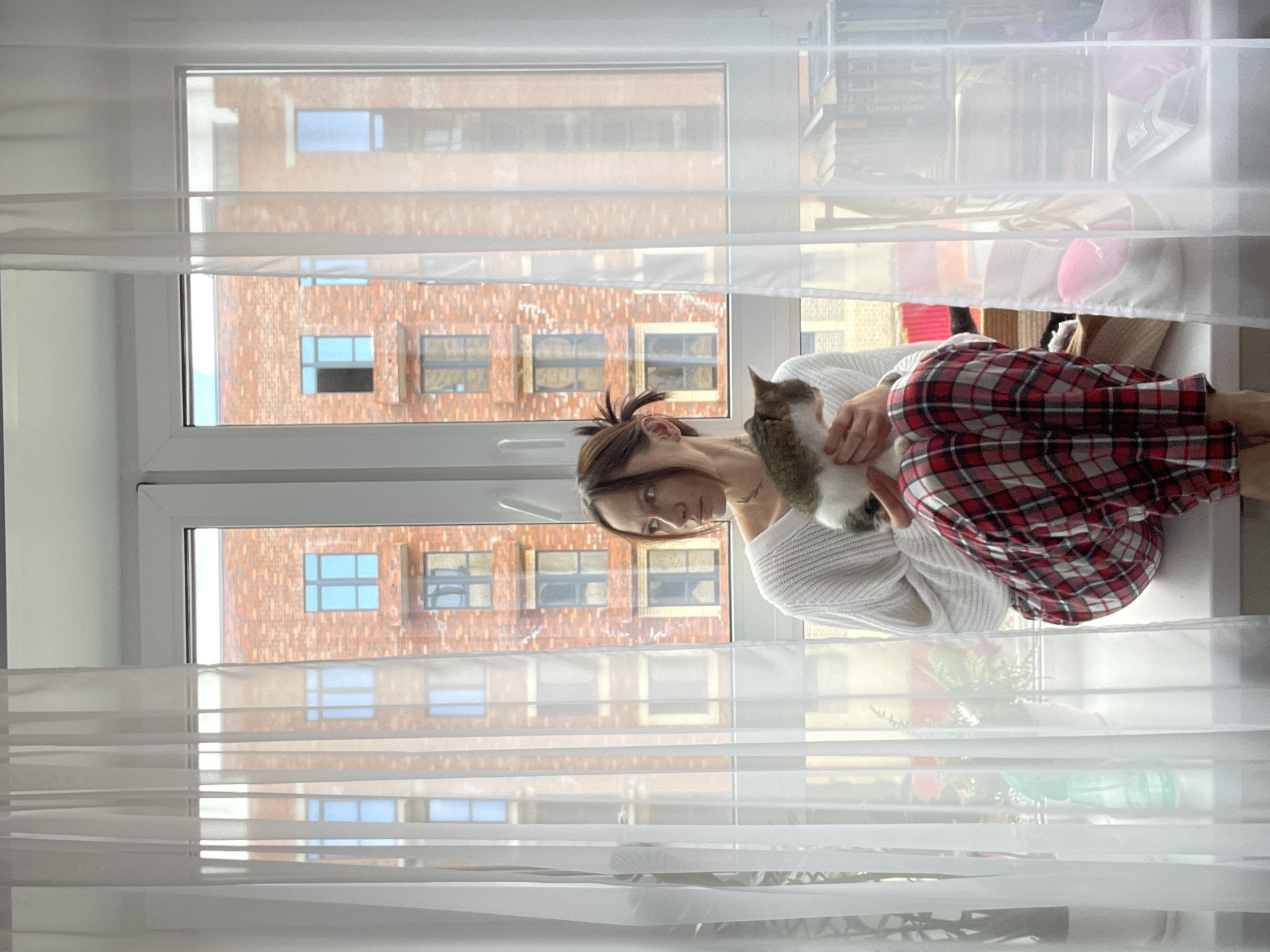 A woman in a white sweater and red-and-black plaid pants sits on a window seat, holding a cat. They are inside a room in Pervomaysky, Russia, with a window showing a brick apartment building across the street. The window is covered by white blinds. Other items visible are plants, a pillow, and books.