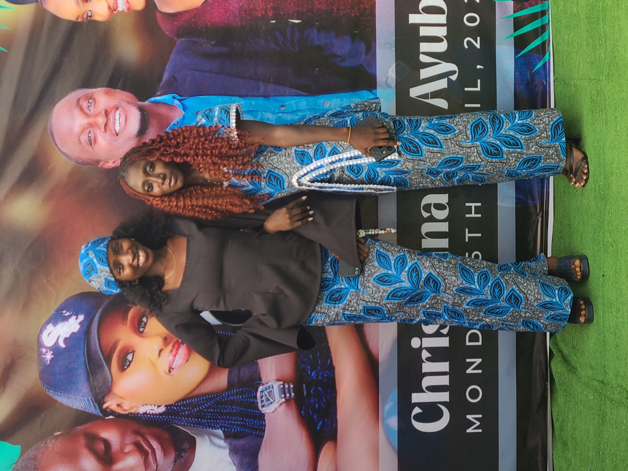 Two women stand in front of a banner featuring several portraits. The woman on the left wears a dark-colored top, patterned blue and grey pants, a blue head wrap, and black sandals. The woman on the right wears a patterned blue and grey outfit with a white-beaded neckline and black sandals.
The banner behind them has portraits of multiple individuals. Text on the banner reads "Chrina" and "Ayub". Another line reads "MOND" and "IL, 202". A patch of green lawn is visible at the bottom right corner of the image.
It is unknown whether this image was taken in Jos, Nigeria or elsewhere.