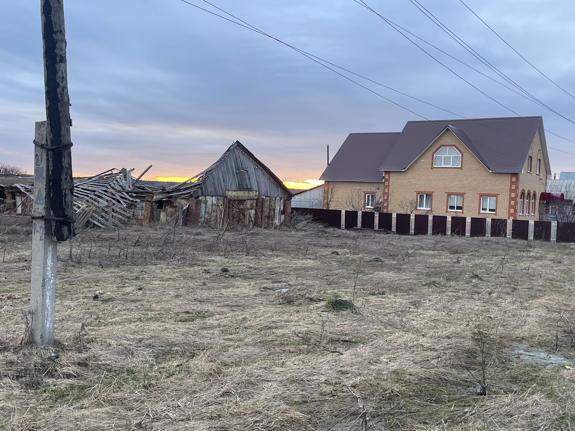 The image depicts a rural or semi-rural scene in Новая Шемурша, Russia. In the foreground, dry, brown grass covers the ground. A concrete utility pole stands on the left side of the frame. Behind it, a dilapidated wooden structure is partially collapsed. To the right of the dilapidated structure, a newer, two-story brick house with a brown roof and a dark-colored fence is present. Overhead, power lines stretch across the sky. The sky is overcast with a hint of orange light near the horizon, suggesting sunrise or sunset. No people are visible in the image.