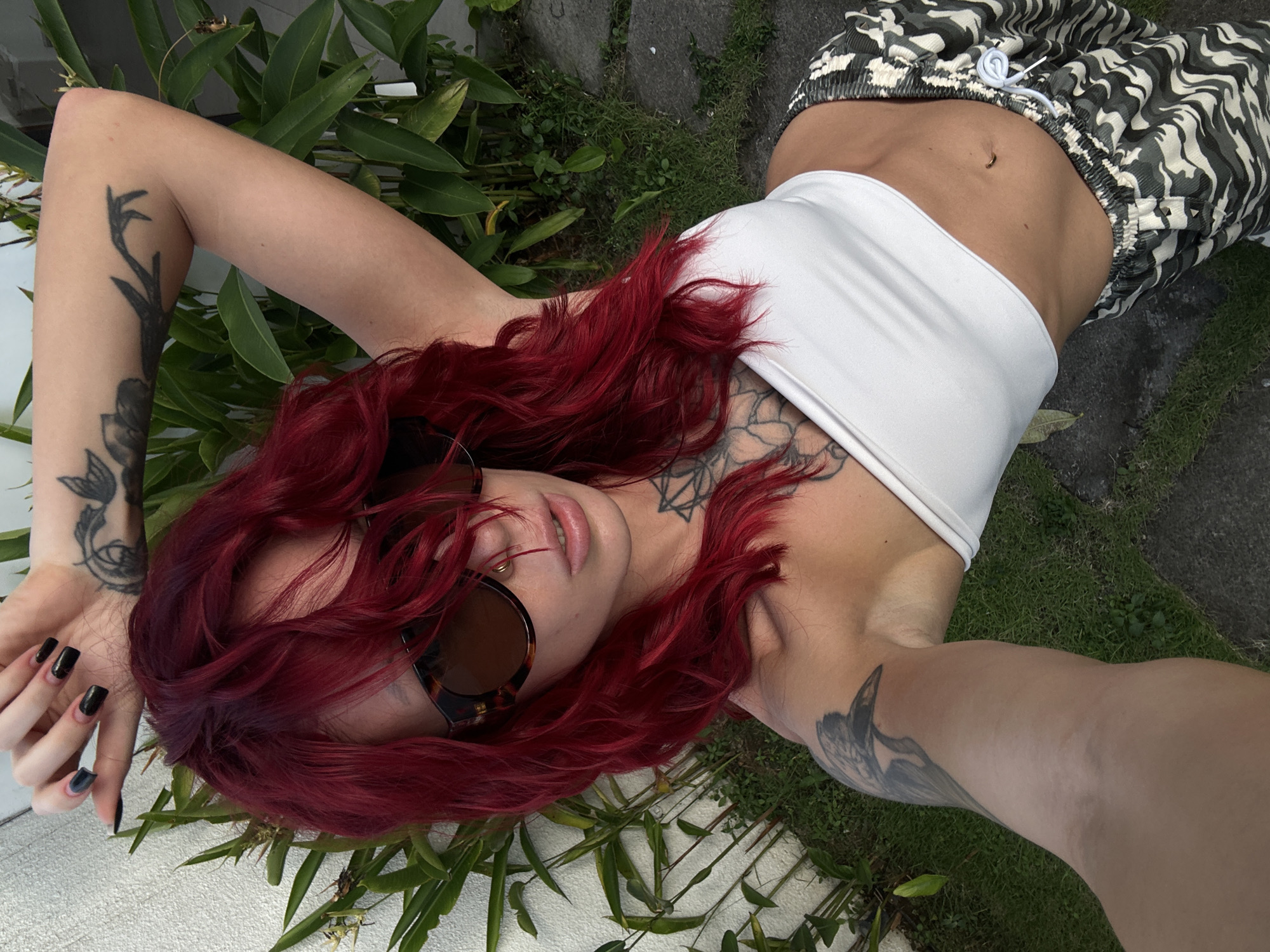 A selfie taken outdoors shows a woman with bright red hair, wearing oversized tortoiseshell sunglasses, a white bandeau top, and camouflage-patterned pants. She has several tattoos visible on her arms and chest. Her left arm is raised behind her head, and her right arm is extended forward, holding the camera. The background includes lush green foliage, a white wall, and a stone pathway. The lighting suggests it is daytime. The location is specified as North Kuta, Indonesia.