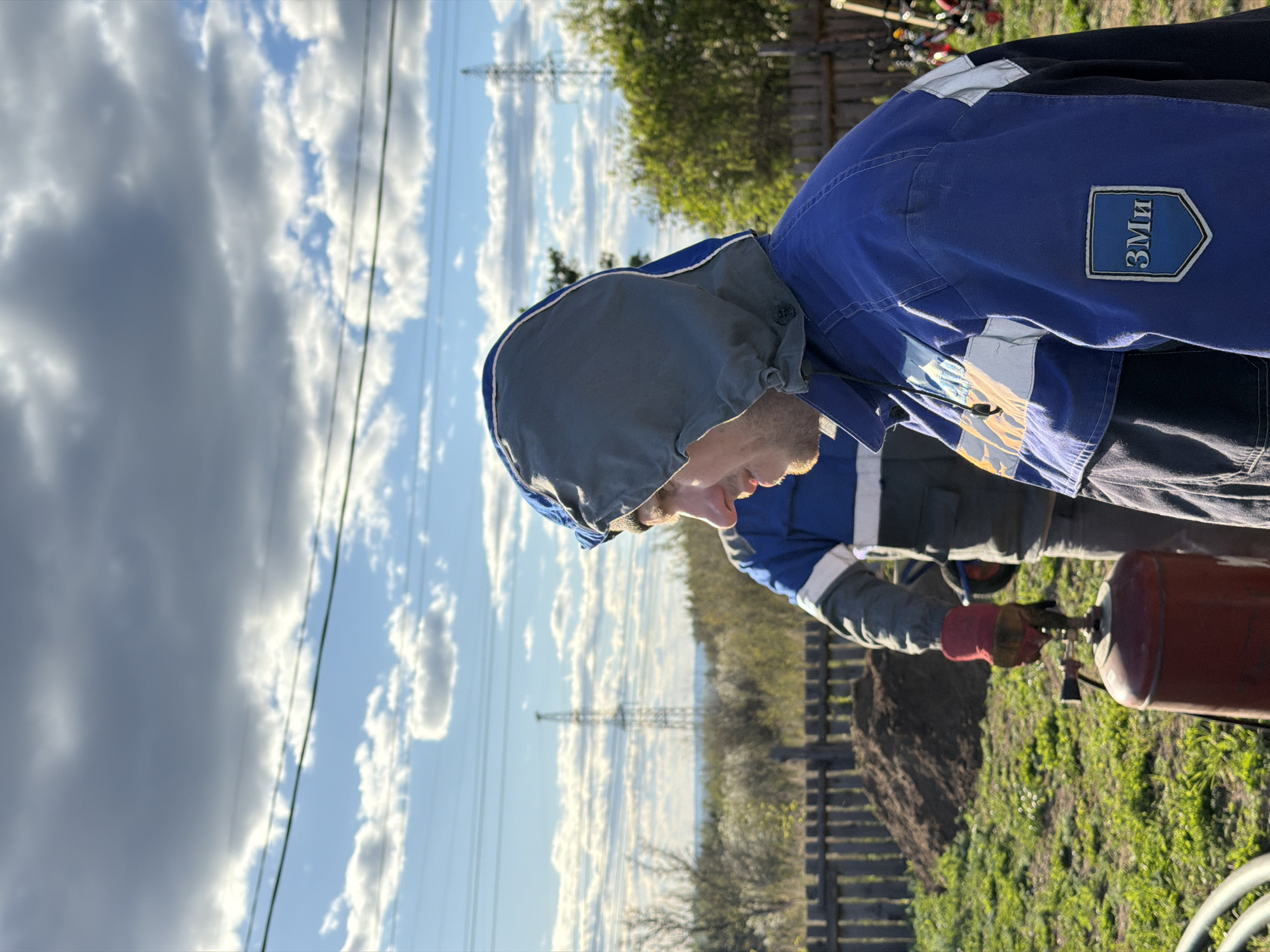 A man in a blue jacket with "3Mn" patch, wearing a gray hood and work gloves, is operating a red propane tank outside, possibly near the city of Chaikovsky, Russia. Background shows power lines, partially cloudy sky, wooden fence, and sparse greenery. The image is rotated 90 degrees clockwise.