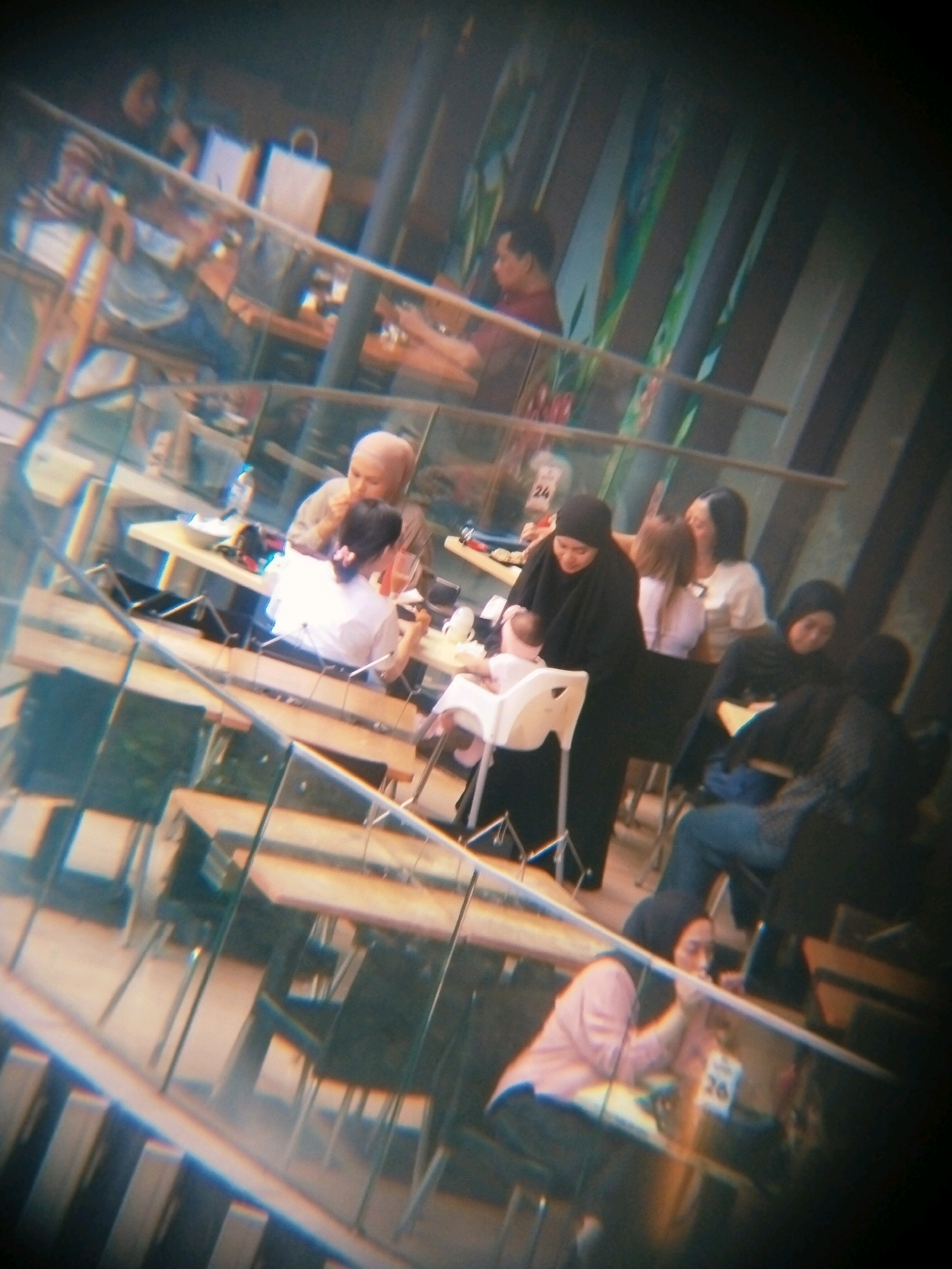 The image depicts a group of people seated at tables in what appears to be an indoor dining area, possibly a cafe or restaurant. The scene is viewed through a distorted lens, creating a prismatic effect that blurs the details.
In the foreground, a person wearing a pink hijab and a pink top is seated at a table, leaning forward and looking down. To their right, a person wearing a dark hijab and a dark top is seated, facing away from the camera. Further in the background, several other individuals are visible, some seated at tables and others standing. One group appears to be a woman in a hijab holding a baby in a high chair. Another individual, a man with a bald head, is seated at a table, looking to his left. In the distance, a person in a red shirt is visible, also seated at a table. The environment is furnished with wooden tables and chairs, and a railing is visible in the upper portion of the image. The overall lighting is dim.
Given the context of Surabaya, Indonesia, this scene is likely a social gathering at a public eating establishment. The presence of hijabs suggests a Muslim-majority population, consistent with Indonesia. The casual attire and the presence of a baby indicate a relaxed, possibly family-oriented setting. The distorted perspective suggests the photograph may have been taken through a reflective surface or with a specialized lens, potentially at an event or a public space where such artistic or unconventional photographic methods are employed.