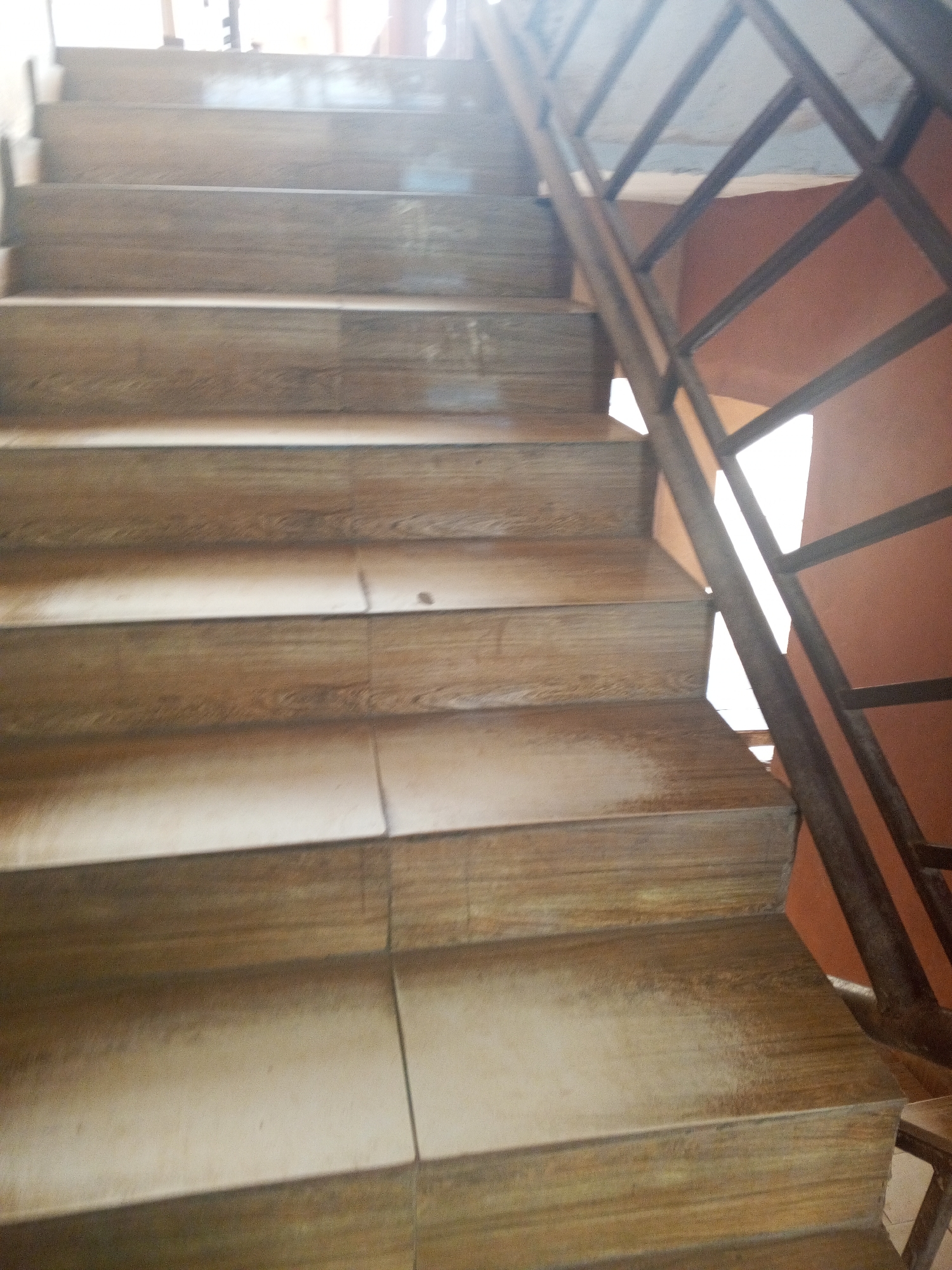 A close-up, eye-level perspective shot captures a set of indoor stairs with a metal railing in Abuja, Nigeria. The stairs appear to be made of wooden planks or tiles, showcasing a natural wood grain pattern in various shades of brown. The steps are evenly spaced and create a sense of depth as they ascend towards the top of the image. A dark metal railing, composed of vertical bars and diagonal supports forming a diamond pattern, runs along the right side of the staircase, indicating it's a functional safety feature. The background is a warm, reddish-brown wall, and the overall lighting suggests an indoor setting, possibly illuminated by natural light filtering in from an opening or window at the top of the stairs. The image is focused on the texture and construction of the staircase, highlighting its simple yet sturdy design.
