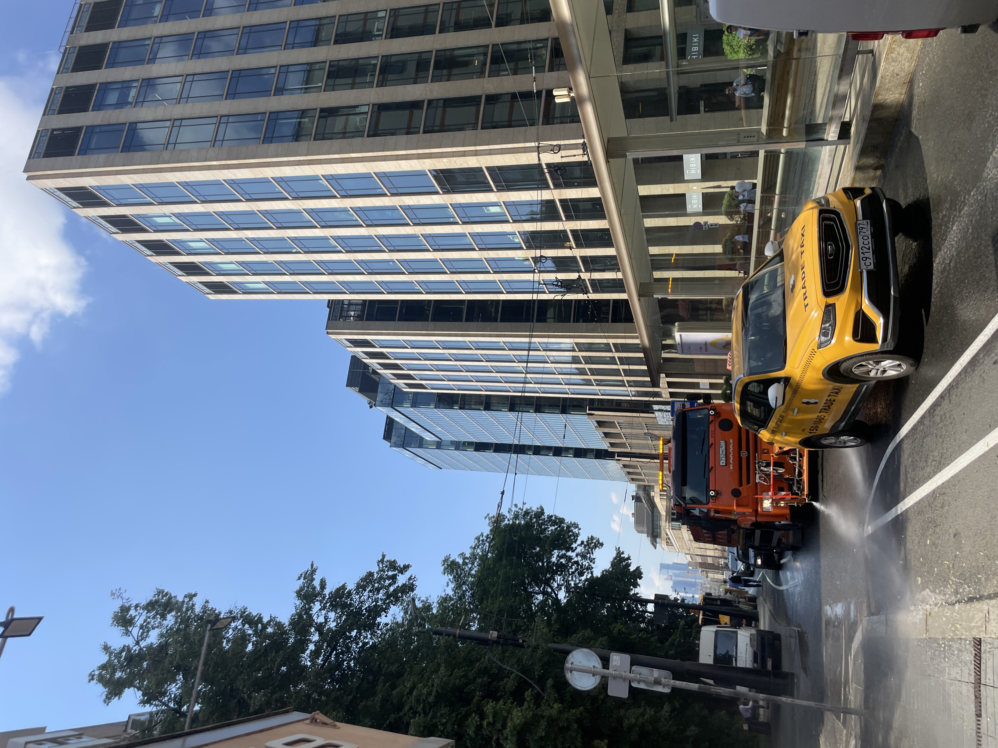In Moscow, Russia, on a bright, sunny day, a street cleaning truck sprays water onto the asphalt. The truck, an orange Kamaz with the number 2123 on its rear, is positioned in the middle of the street, facing left. A yellow Geely Coolray taxi, marked "TRADE TAXI" and with license plate C912CO 797, is driving past the cleaning truck, also heading left. The taxi is slightly wet from the spray.
The scene is bordered by a tree-lined sidewalk on the left and modern glass-fronted buildings on the right. The buildings feature a light-colored facade and dark-tinted windows. A covered walkway or bus stop with glass panels runs along the front of the buildings.
In the background, more buildings and a clear blue sky are visible. A white van is parked on the left side of the street, and a few people are seen walking and sitting on a bench near the van. Power lines are strung across the scene above the street. The overall environment suggests an urban setting with active street maintenance.